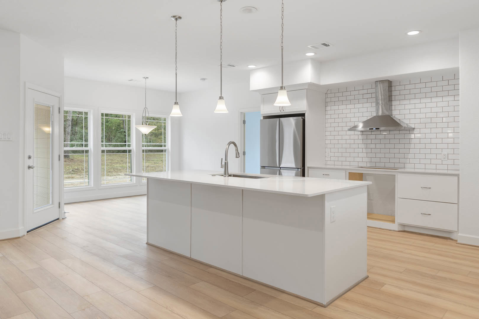 White kitchen with wood flooring, large central island featuring stone countertop, stainless steel exhaust hood above stove, white cabinetry, tile backsplash, window overlooking