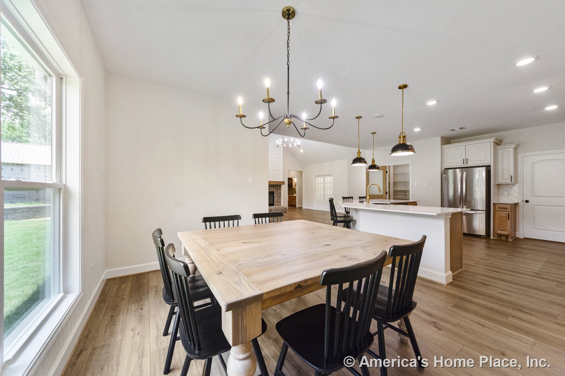 Open dining and kitchen area with wood plank flooring, white cabinetry, kitchen island with pendant lights, black chandelier above dining space, large window with decorative trim