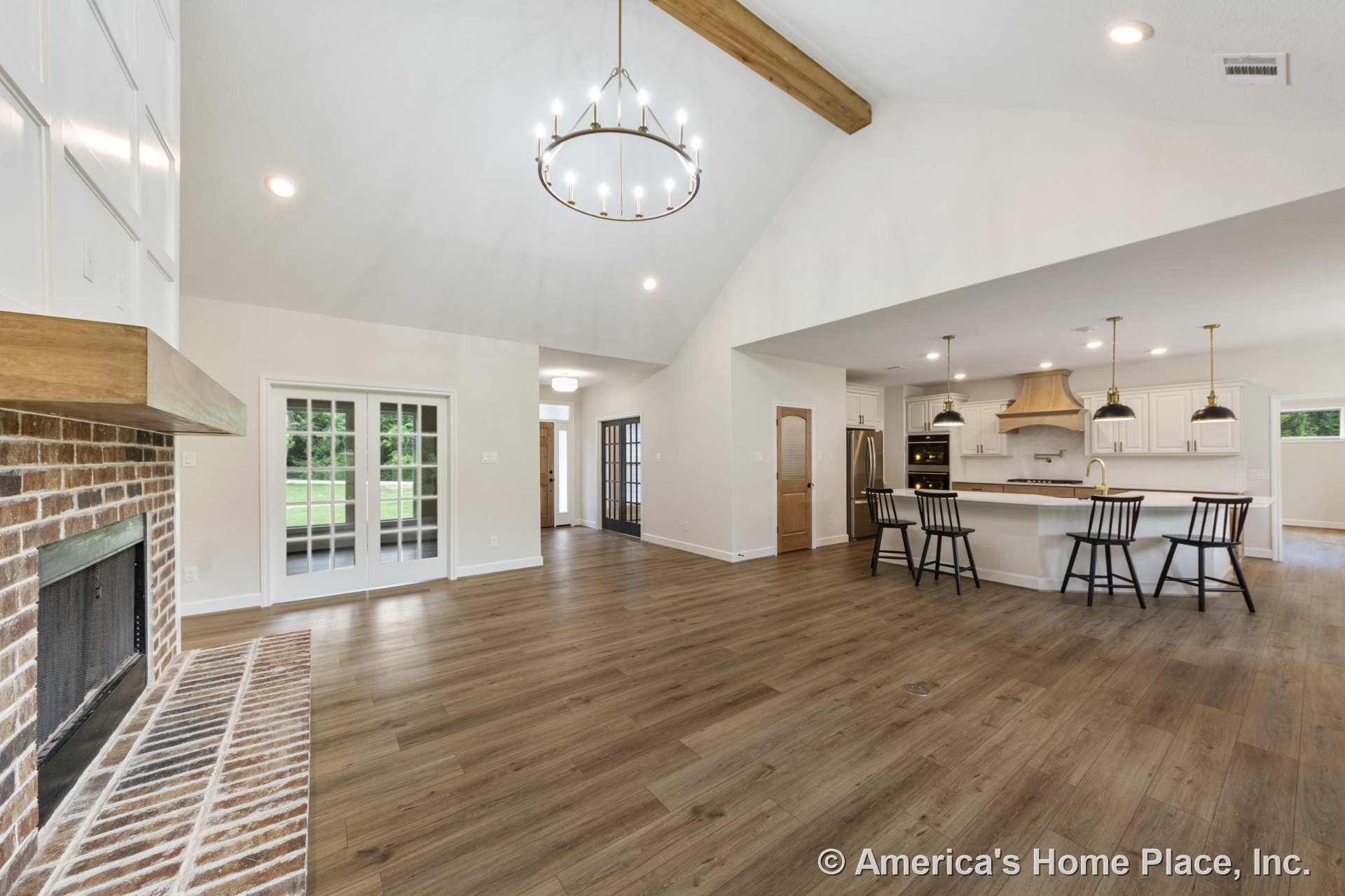 Open-concept living area with brick fireplace and wood mantel, vaulted ceiling with exposed wood beam, large chandelier, adjoining kitchen featuring white cabinetry, central