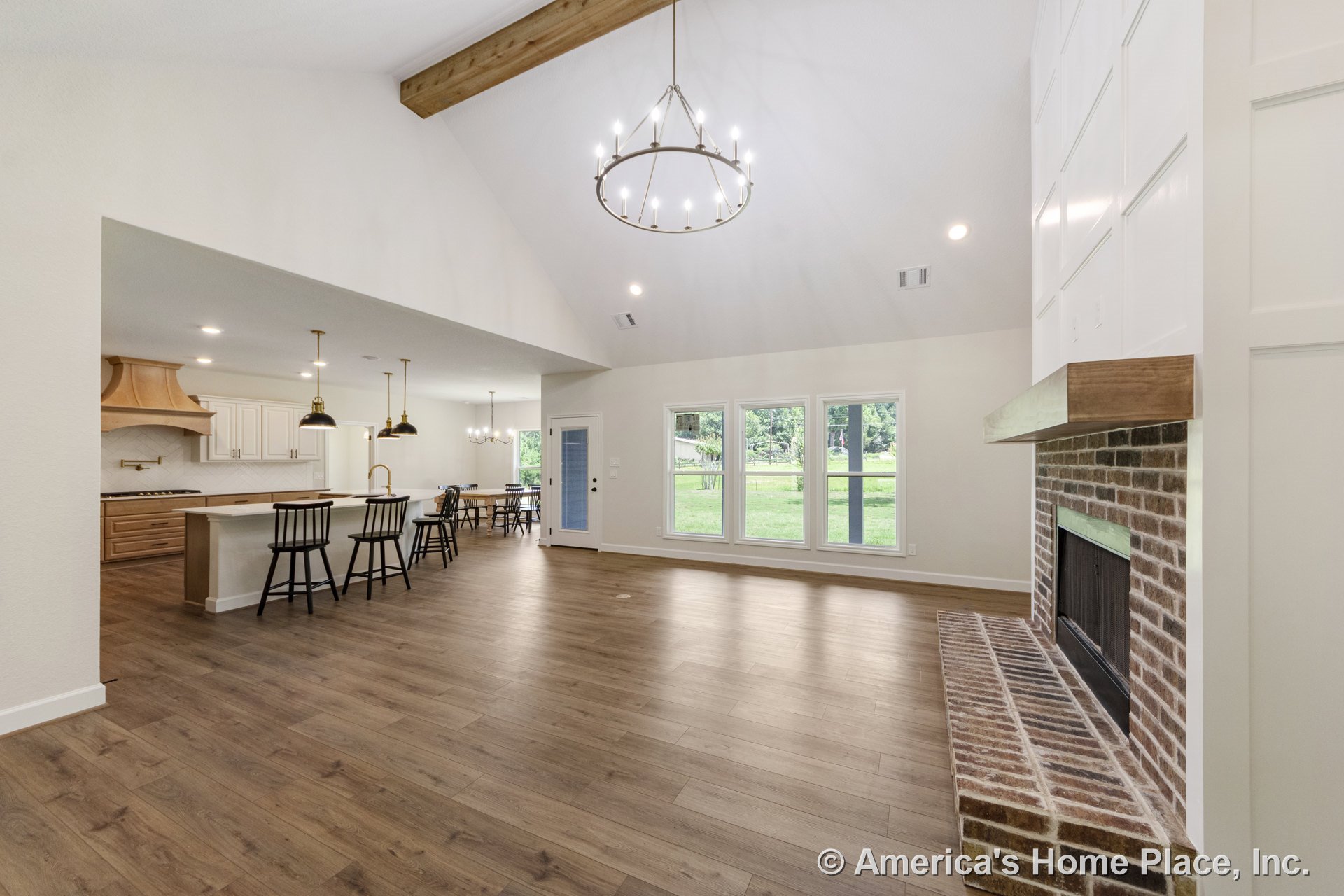 Brick fireplace with wood mantel, vaulted ceiling with exposed beam and chandelier, wide plank wood flooring, large windows with white trim, open kitchen featuring island with