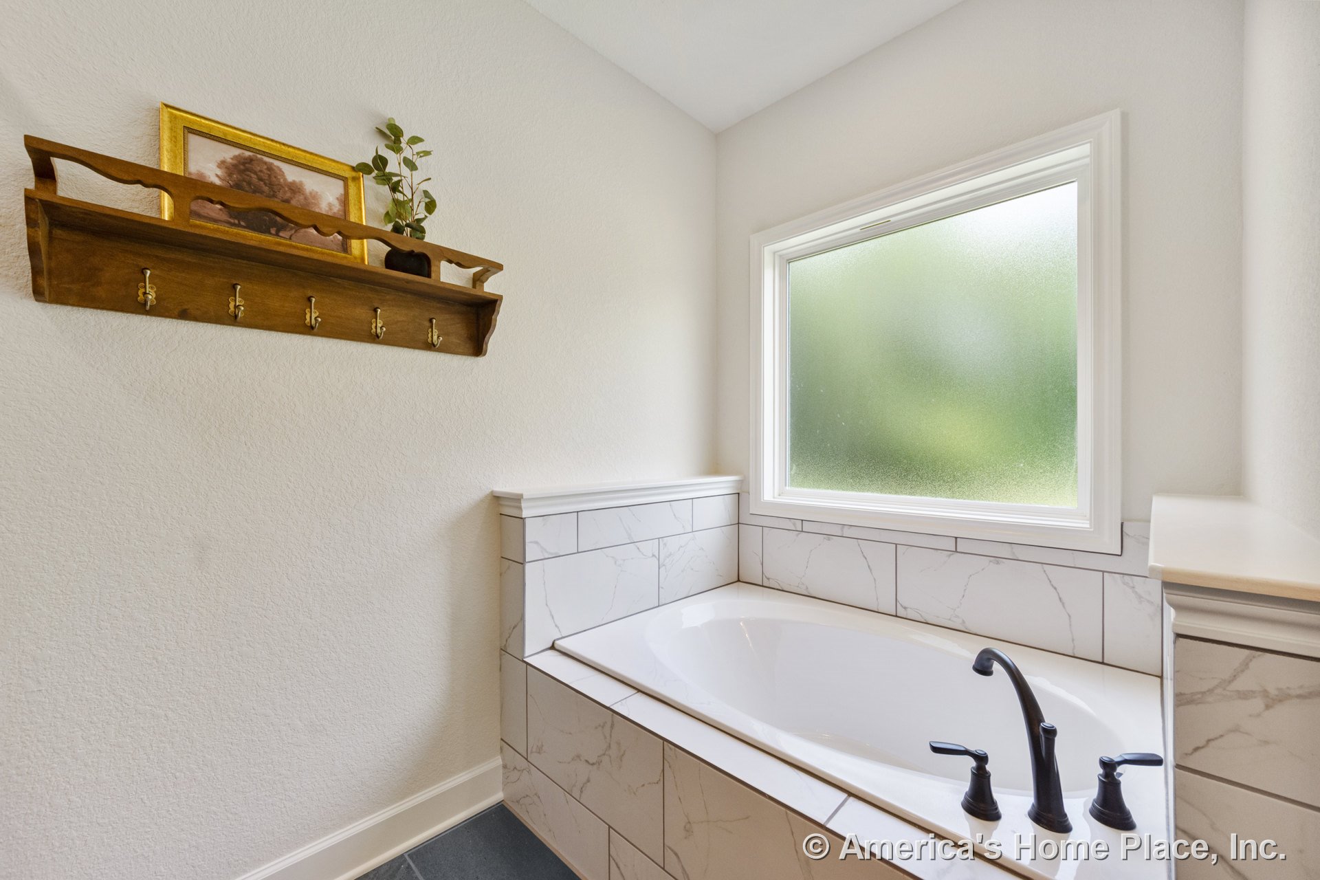Built-in soaking tub with black fixtures and marble-look tile surround beneath a frosted window with white trim; wall-mounted wood shelf with hooks, light textured wall finish, and