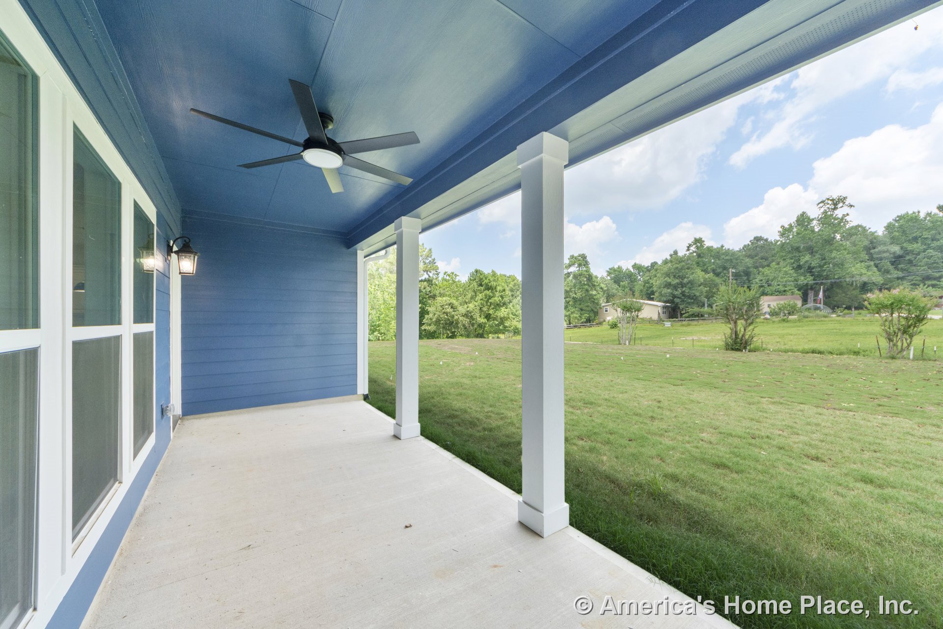 Covered porch with blue siding, white trim, concrete flooring, ceiling fan, exterior windows, and white columns.