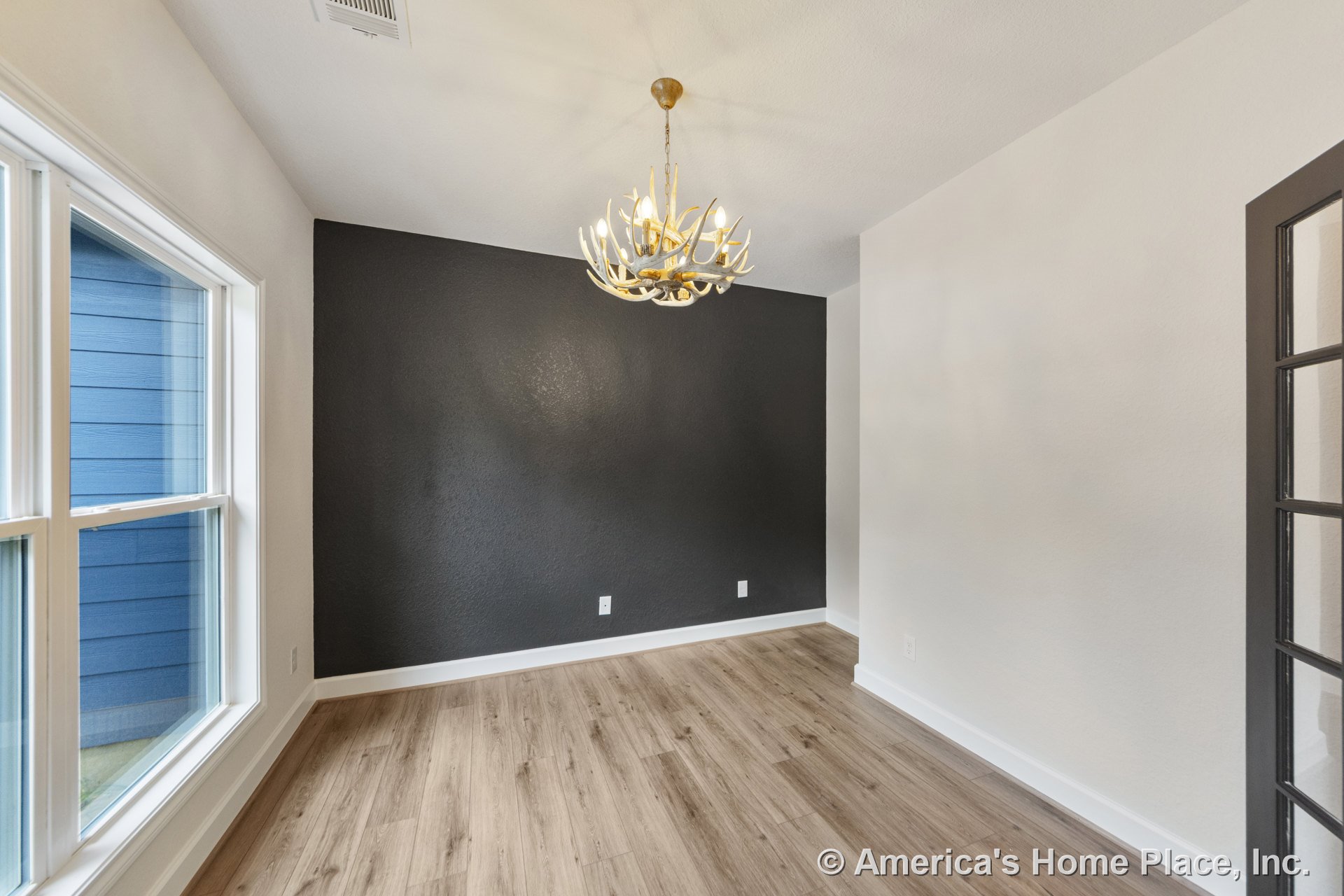 Dining room with black accent wall, antler chandelier, wood-look plank flooring, large windows, white trim, and glass-paneled door allowing natural light.