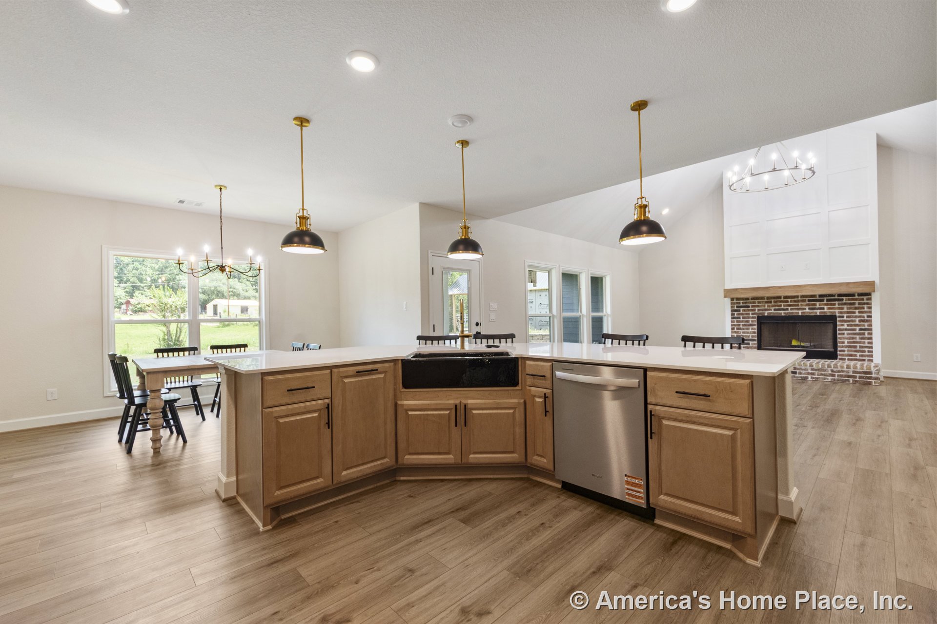 Open concept kitchen with light wood cabinetry, quartz countertops, black farmhouse sink, and pendant lighting, overlooking living area with brick fireplace, wide plank flooring