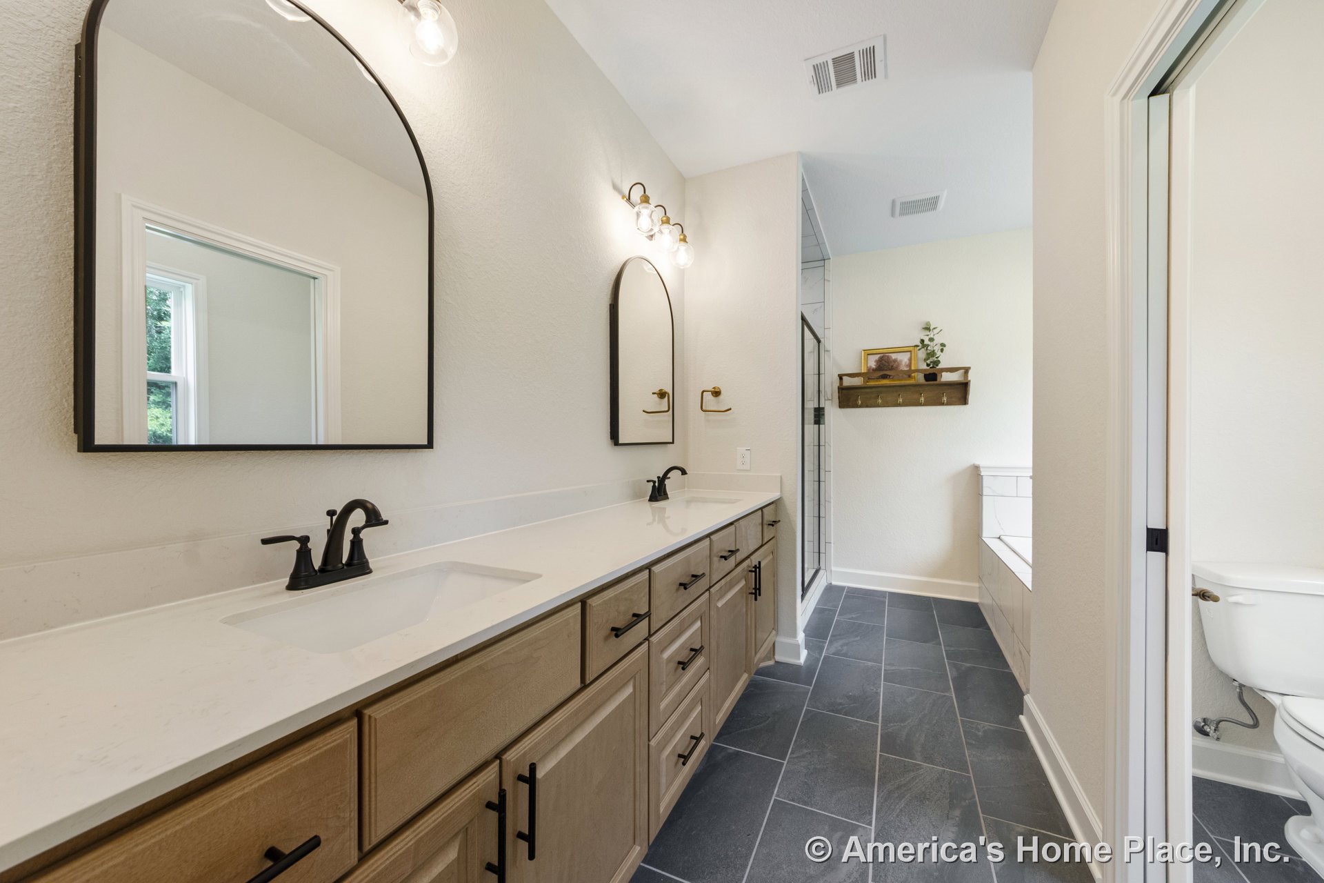 Double vanity with light wood cabinetry, quartz countertops, matte black faucets, arched mirrors, wall-mounted globe sconces, gray tile flooring, glass shower enclosure, built-in