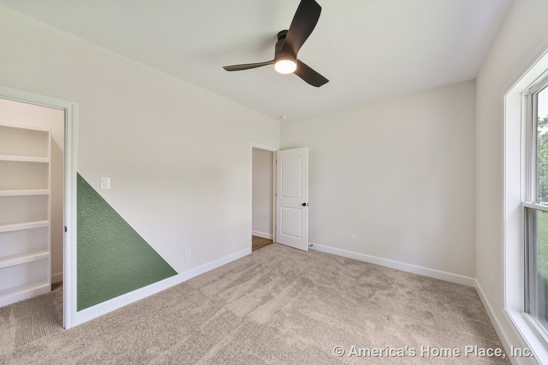 Beige carpeted bedroom with modern ceiling fan, built-in closet shelving, large window with white trim, accent wall paint, and neutral walls.