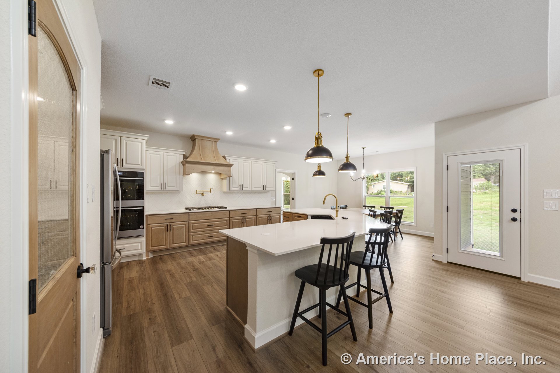 Spacious kitchen with large quartz island, wood cabinetry, custom wood range hood, double wall ovens, white upper cabinets, wide plank wood flooring, gold faucet, pendant and