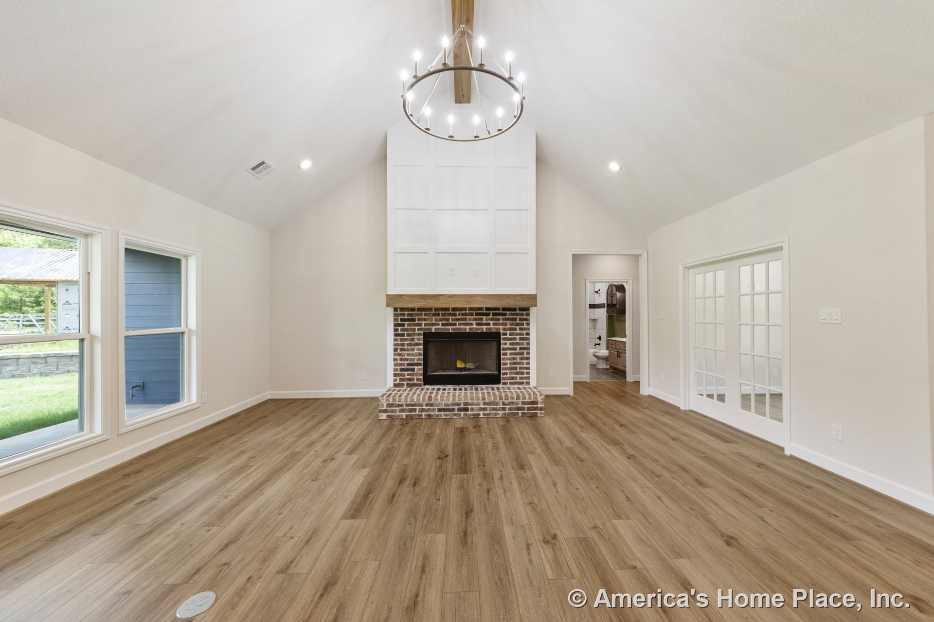 Vaulted ceiling with exposed beam, large brick fireplace topped by wood mantel, wide plank wood flooring, double glass French doors, expansive windows with white trim, recessed