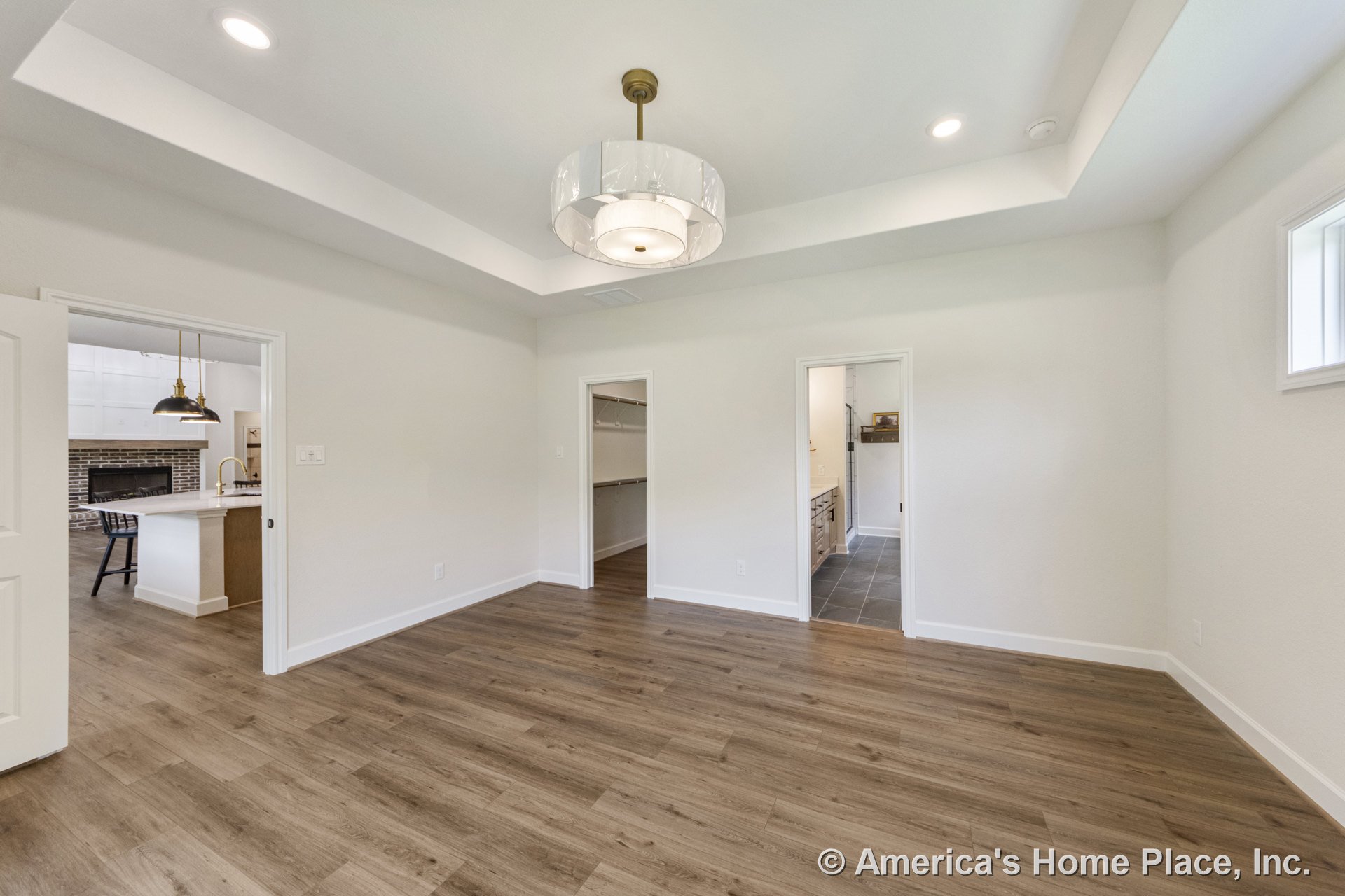 Bedroom with tray ceiling, recessed lights, modern pendant fixture, wood-look plank flooring, white trim, neutral walls, doorway to walk-in closet, and bathroom featuring tile