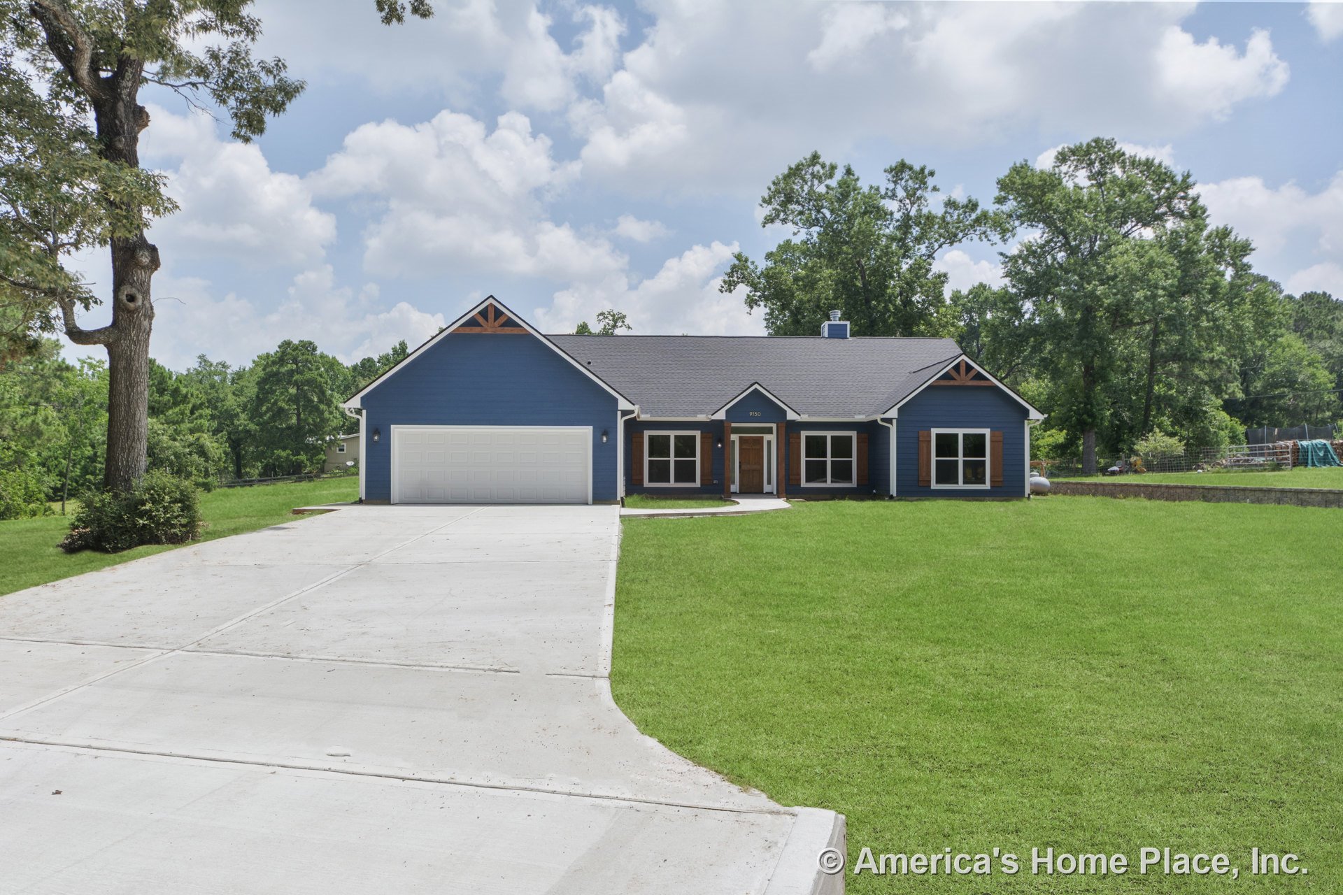 Blue siding exterior with wood trim gable accents, large concrete driveway, white garage door, multiple front windows, covered front entry, and single-story layout with lawn.