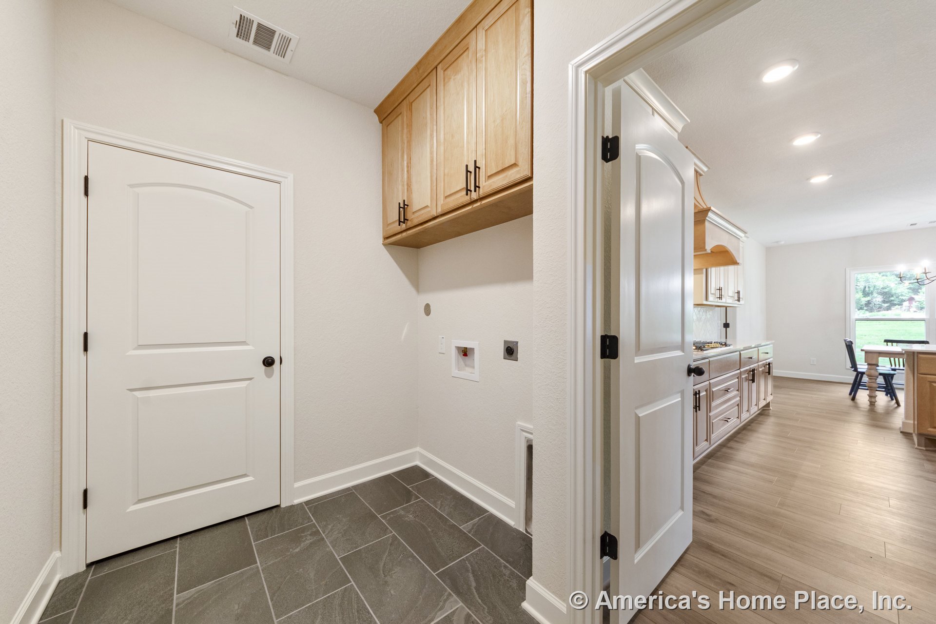Laundry room with built-in natural wood upper cabinets, slate tile flooring, white paneled doors, utility hookups, and recessed ceiling lights; open doorway leads to kitchen with