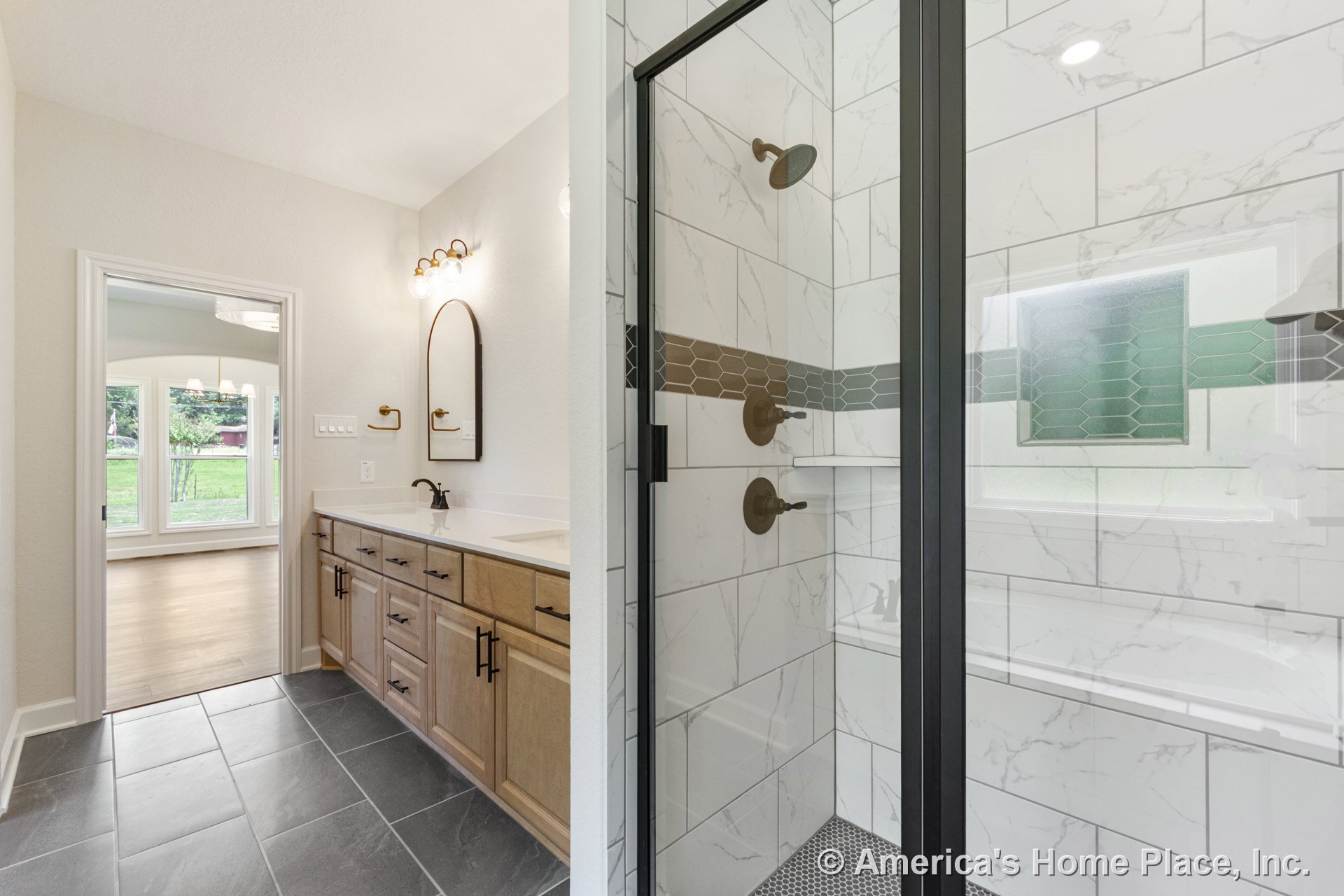Glass-enclosed shower with marble-look tile, double vanity featuring wood cabinetry and black hardware, large bathroom mirror with wall-mounted lighting, dark slate floor tile, and