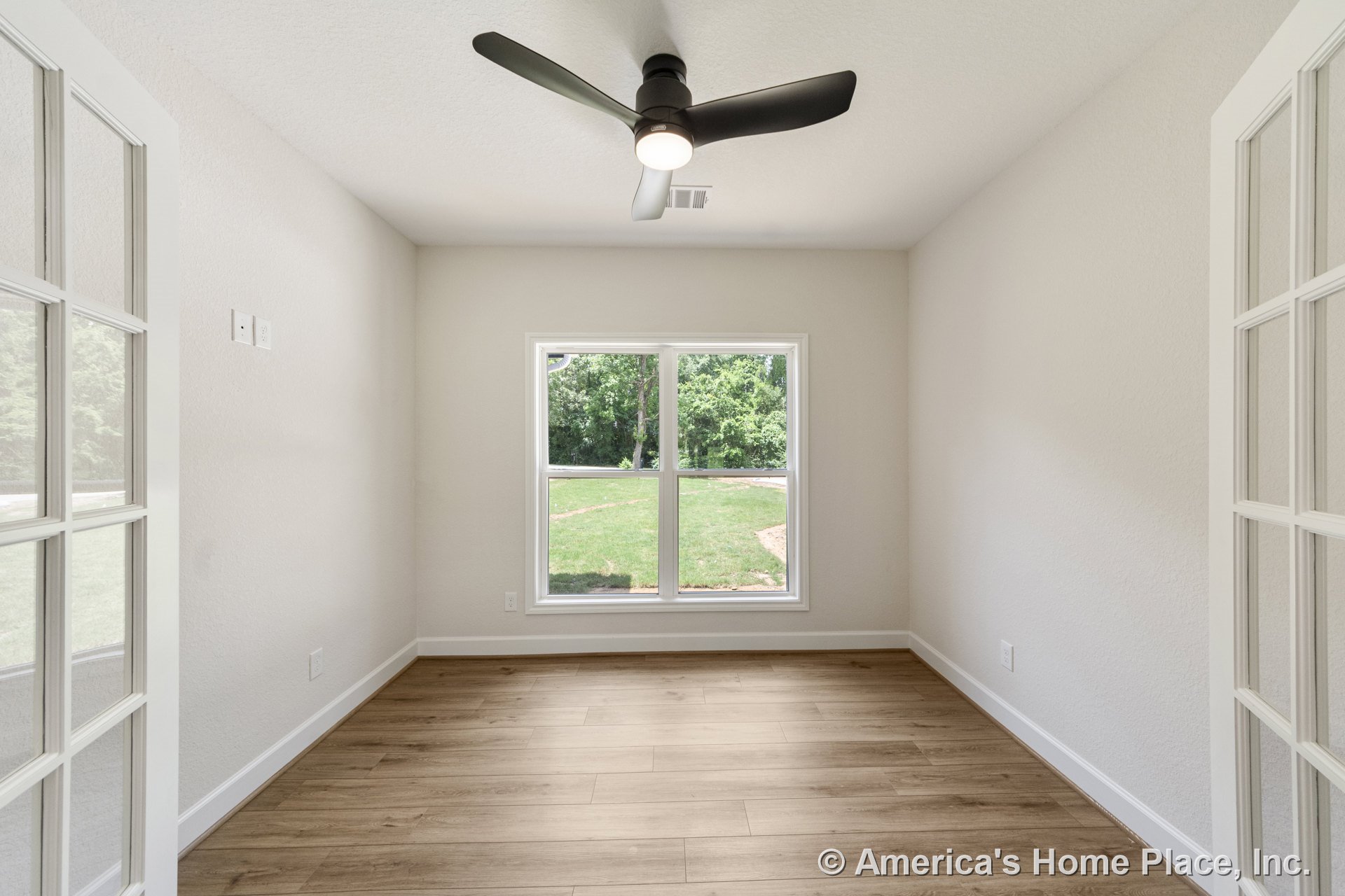 Small room with light wood plank flooring, matte black ceiling fan with integrated light, double French doors, large window with white trim, neutral painted walls, and baseboard