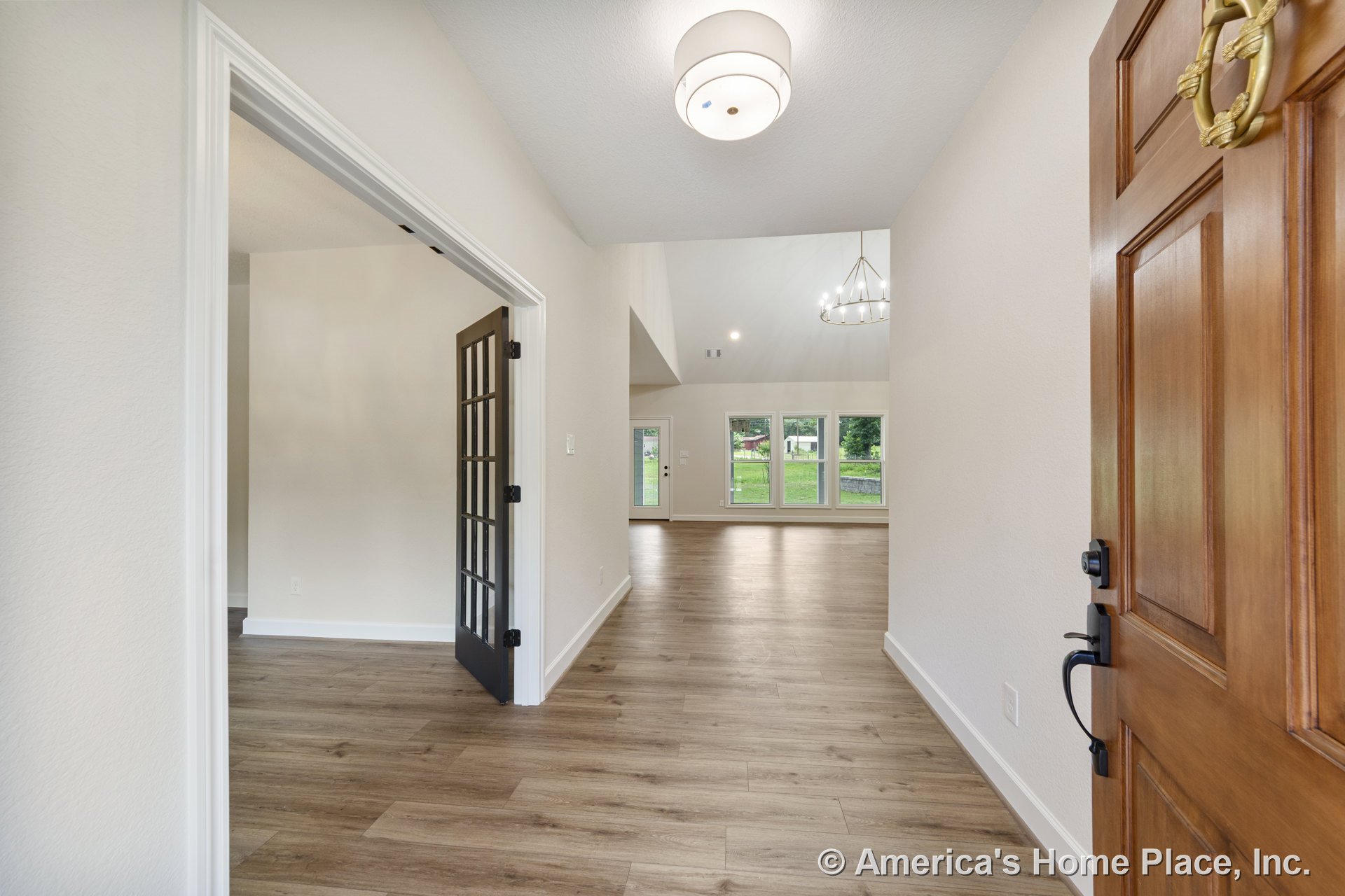 Light wood plank flooring in entryway with white baseboards, modern flush mount ceiling light, black framed glass door, and open view to living area featuring vaulted ceiling