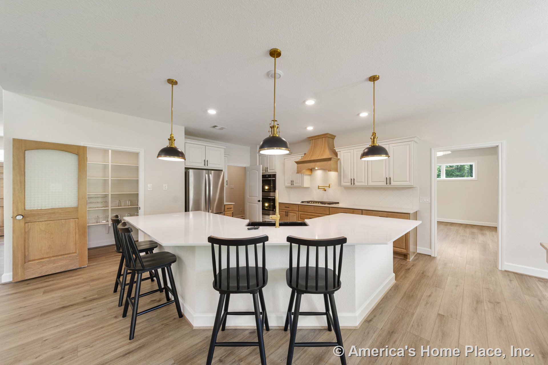 Spacious kitchen with large white quartz island featuring built-in sink, brass pendant lights, wood cabinetry and range hood, stainless steel appliances, white upper cabinets