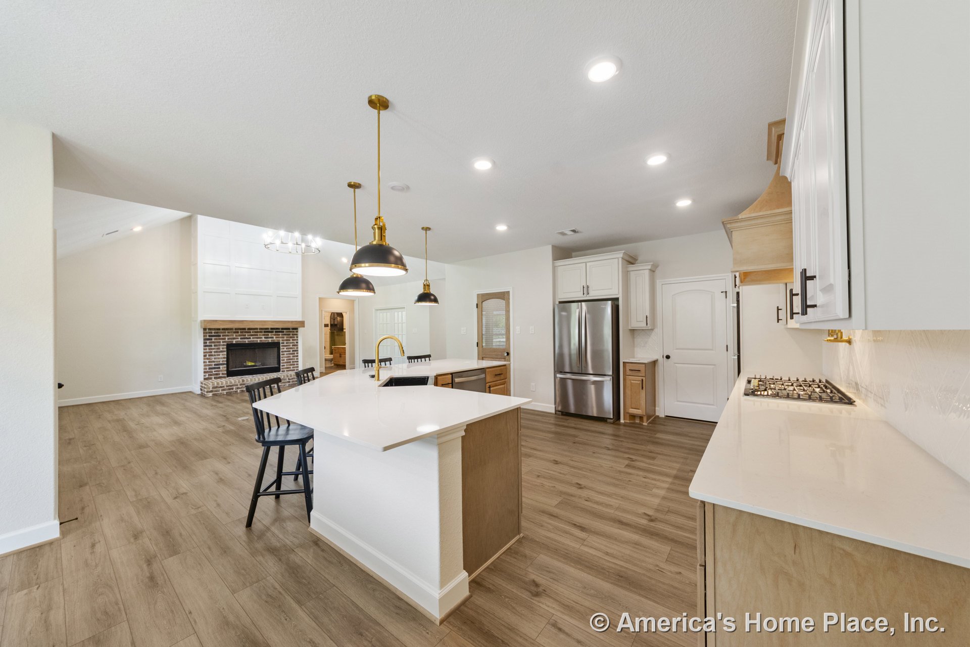 Open kitchen with large quartz island, wood cabinetry, white upper cabinets, stainless refrigerator, pendant and recessed lighting, wood-look flooring, and view into living area