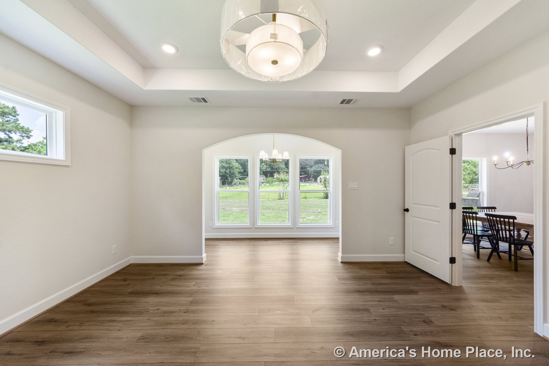 Dining area with wide plank wood flooring, tray ceiling featuring recessed lights and a modern fixture, large arched windows with white trim, open doorway leading to adjacent room