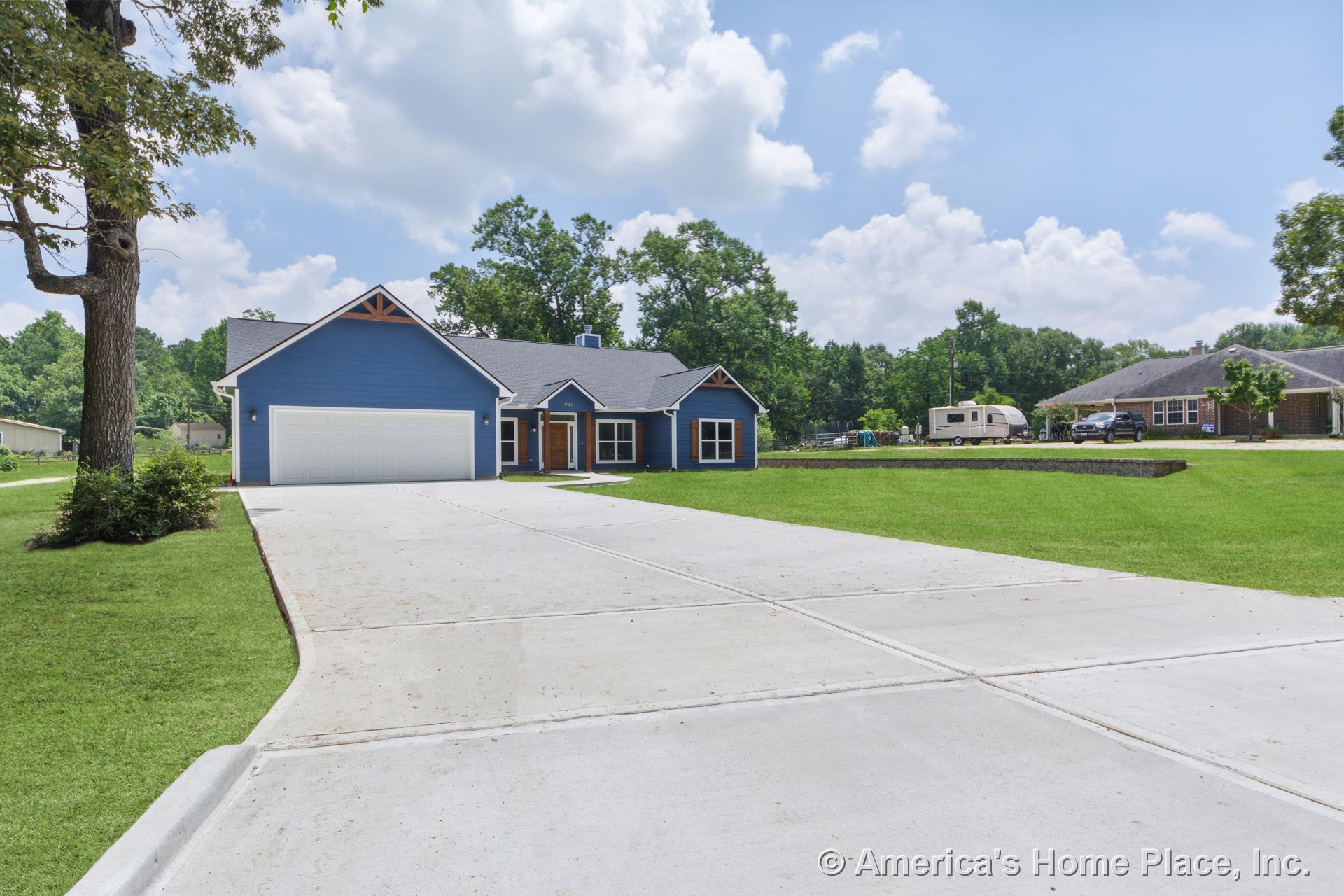 Blue horizontal siding with white window trim, gabled roof featuring wood accents, large concrete driveway leading to a white two-car garage, covered front porch, landscaped front