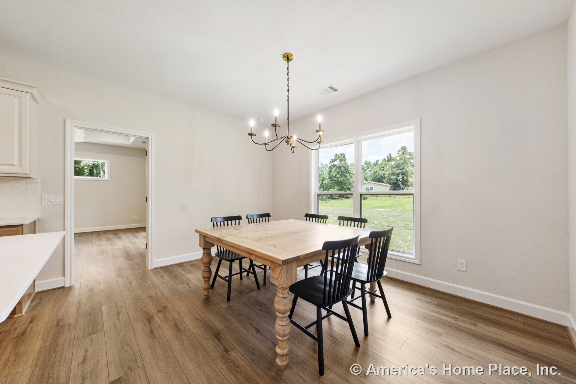 Dining area with large double windows, wood-look plank flooring, white baseboard trim, ceiling-mounted chandelier, open doorway to adjacent room, and neutral wall color.