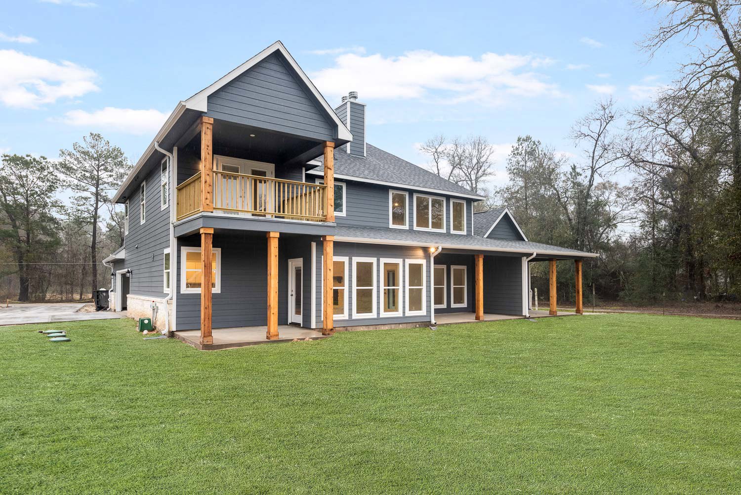 Two-story home with white siding, covered front porch featuring wooden railings, second-floor balcony, manicured green lawn, and tall trees in the background.