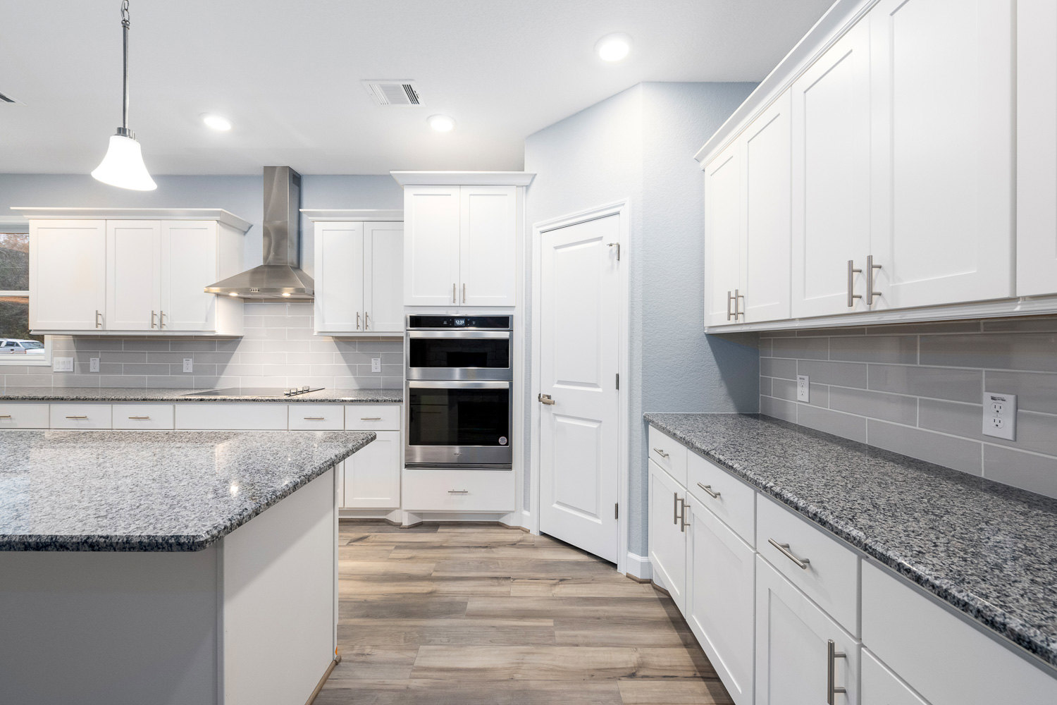 White kitchen with granite countertops, stainless steel oven, gray tile backsplash, white door with silver handle, and outlet on wall.