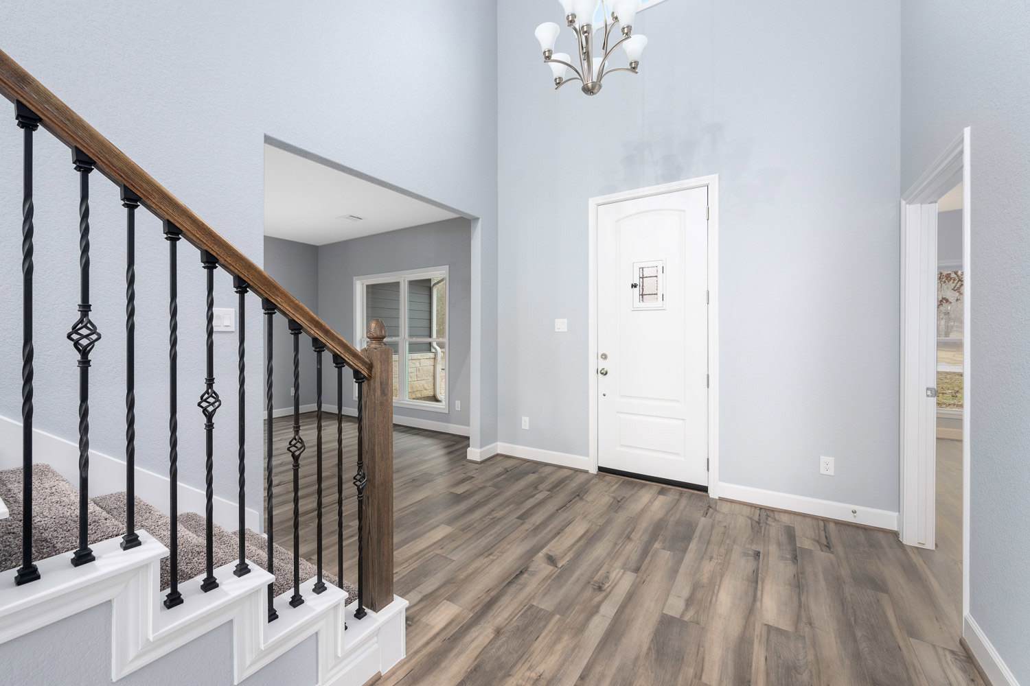 Hallway with light wood flooring, white door featuring a small window, wooden staircase with matching railing, and a modern chandelier overhead