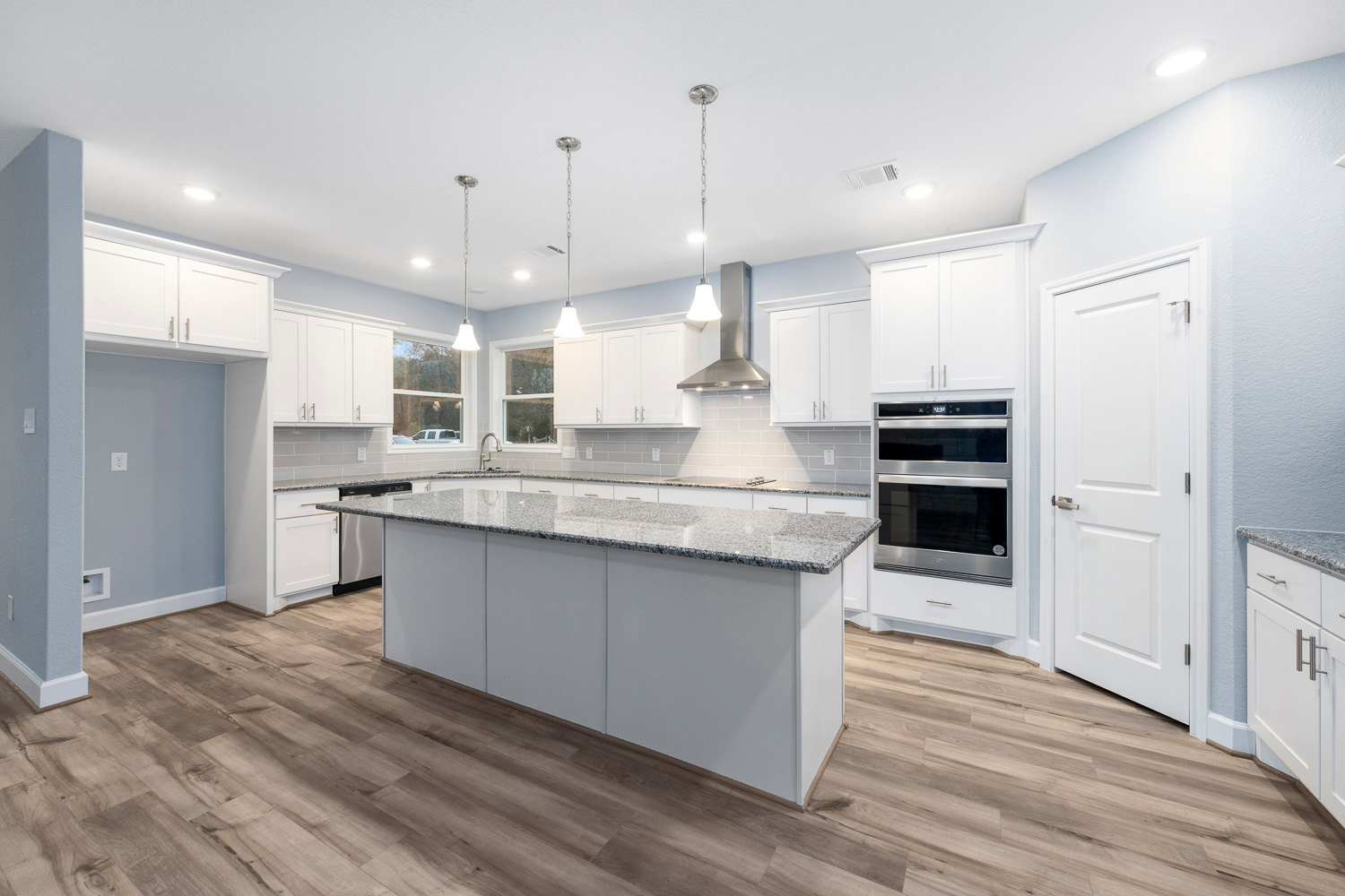 Kitchen featuring white shaker cabinets, wide-plank wood flooring, marble island countertop, stainless steel double oven, undermount sink, and brushed silver hardware