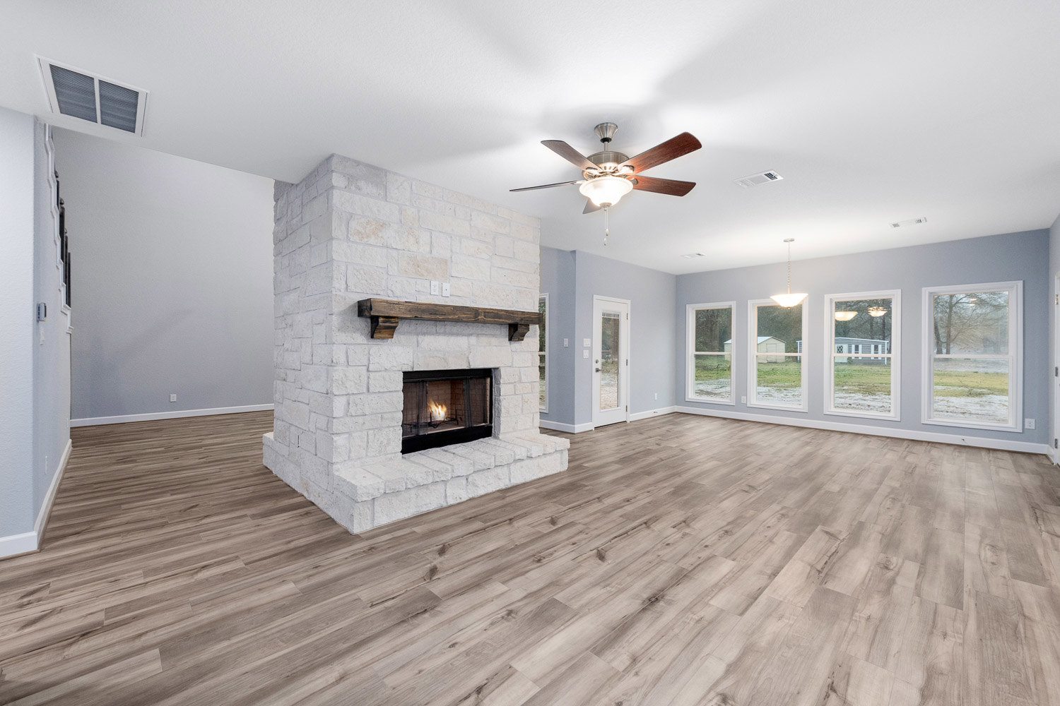 Living room with wood flooring, stone fireplace featuring a wood mantle, ceiling fan with light, and large window overlooking snowy landscape