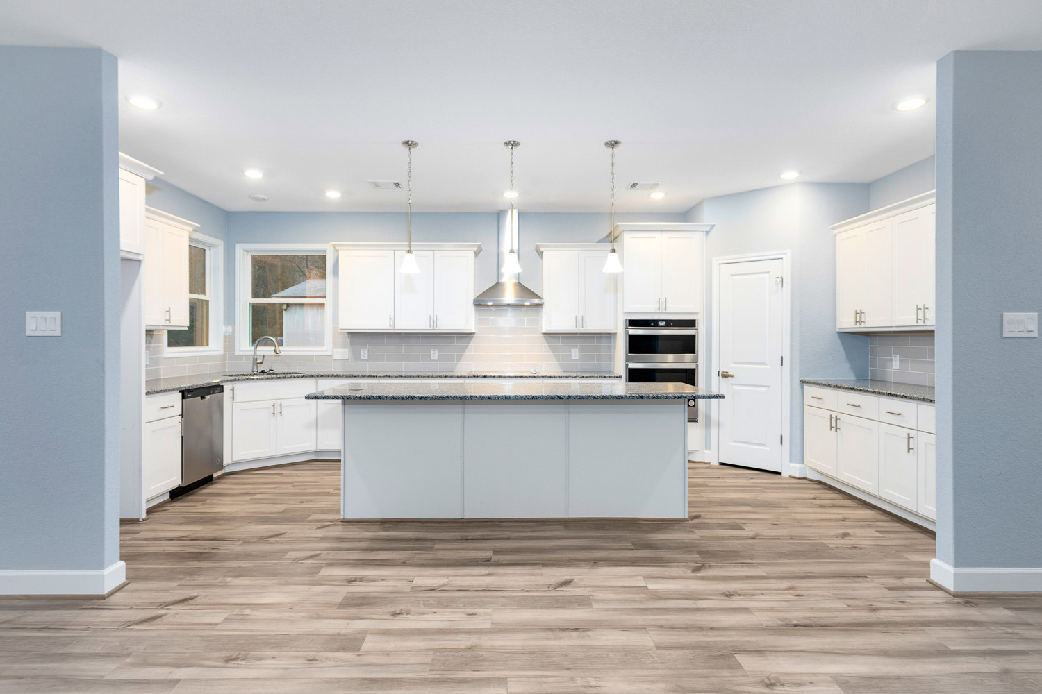 White kitchen cabinets with black-speckled countertops, stainless steel oven with digital display, wood flooring, white door with gold handle, faucet on counter, and white light