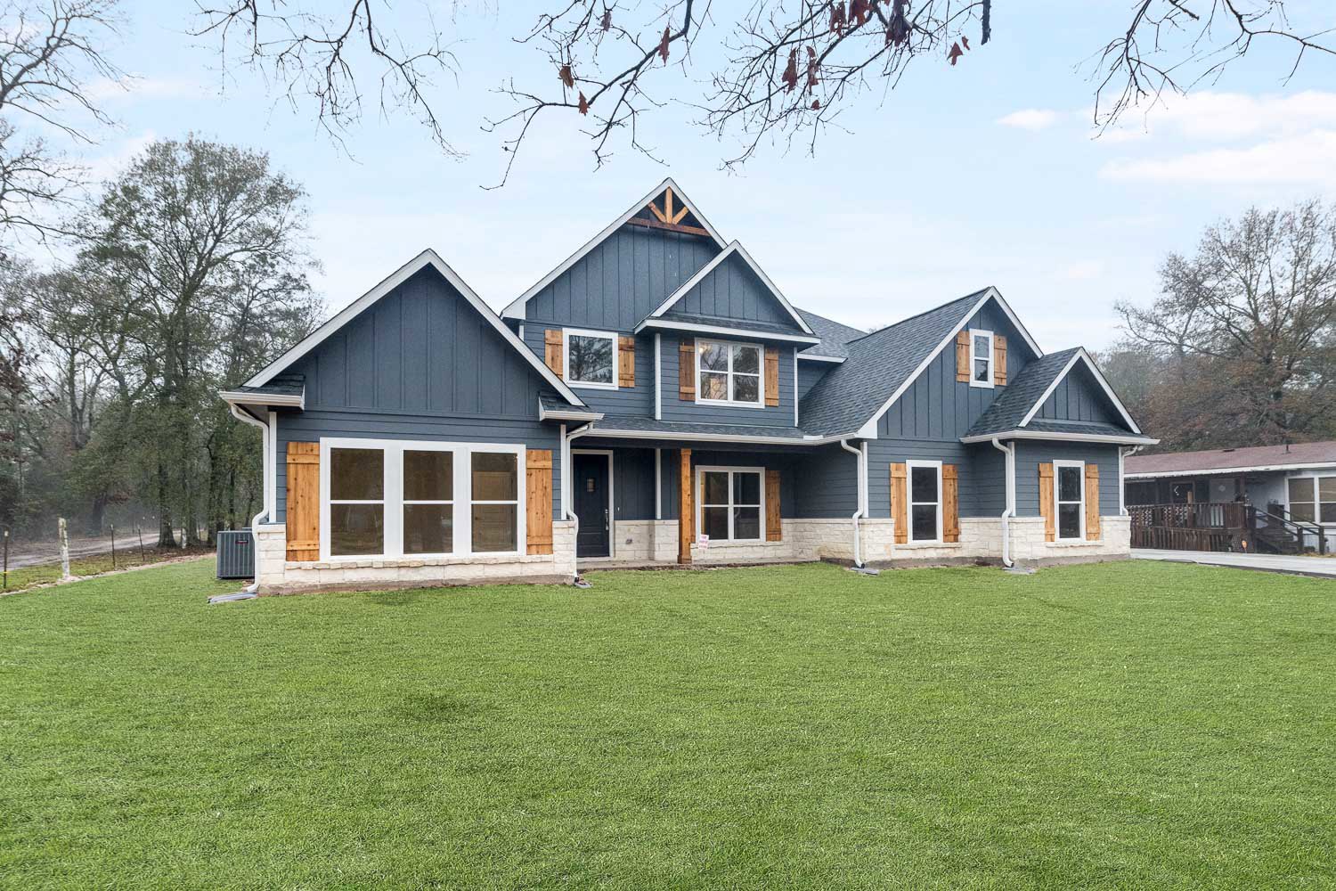 Two-story house with blue roof, brick exterior, large windows, front porch, and green lawn bordered by trees