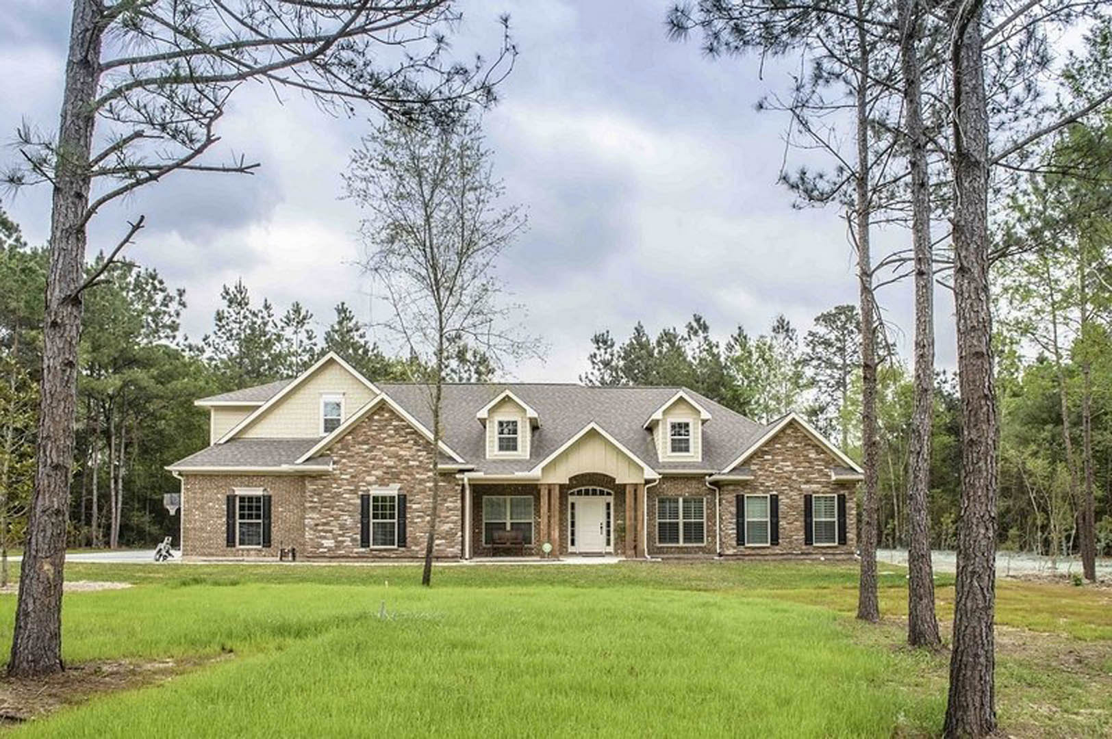 Two-story farmhouse with white siding, white-framed windows, and a glass-paneled front door, surrounded by a manicured green lawn and mature trees under a partly cloudy sky