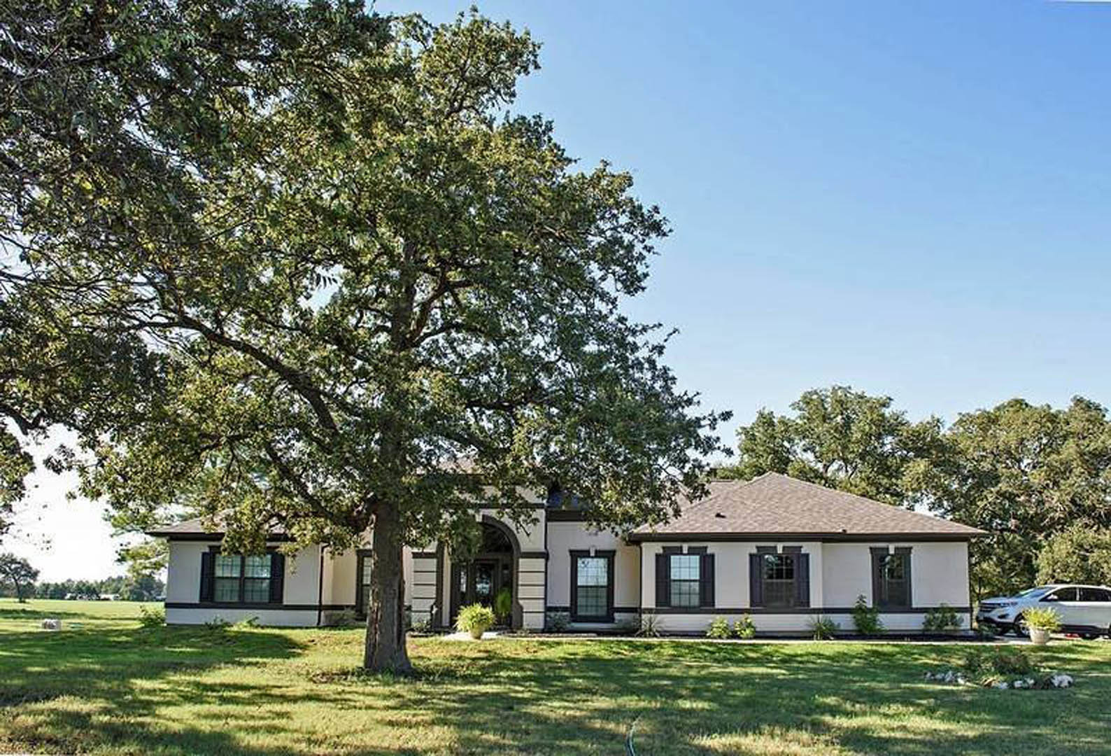 Two-story home with light siding, large grid windows, prominent tree in landscaped front yard, white car parked on driveway, potted green plant near entrance, blue sky overhead