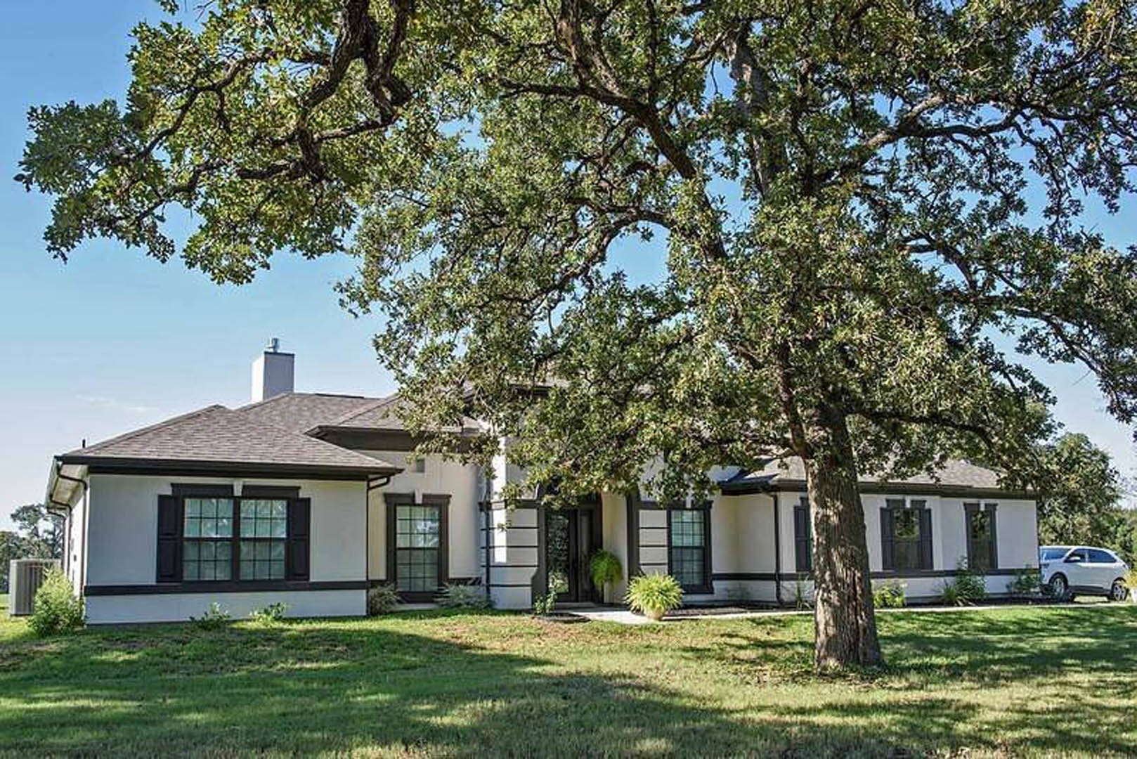 Two-story farmhouse with gray siding, large front lawn, mature trees, white car parked roadside, multi-pane windows, potted green plant near entry, close-up of wooden front door