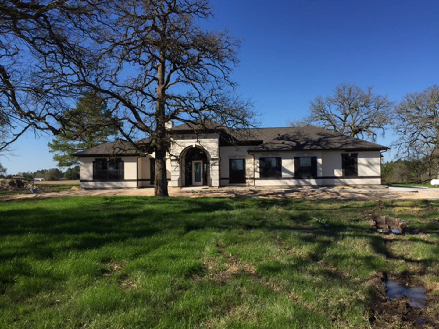 Two-story farmhouse with white front door, gray siding, large windows, and a mature tree in the backyard, set on a grassy lawn under a blue sky
