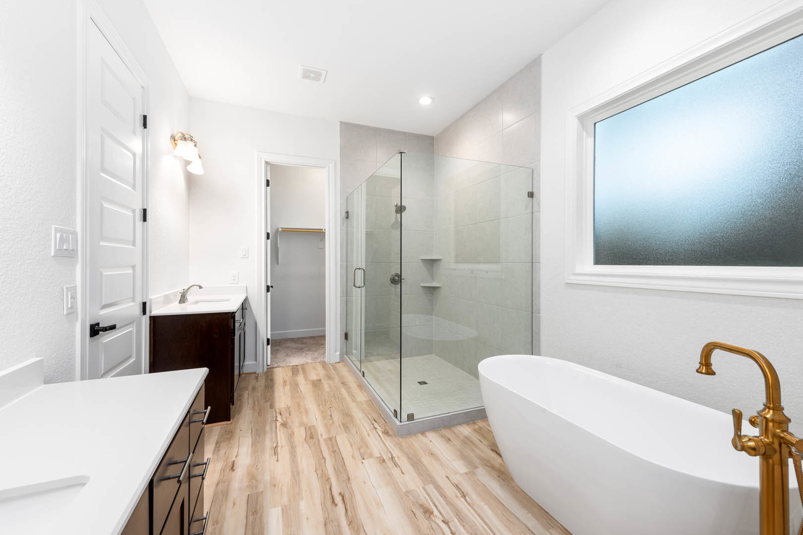 Bathroom featuring a frameless glass shower, freestanding white tub with gold faucet, white countertop, brown vanity cabinet, and tiled floor with a window.