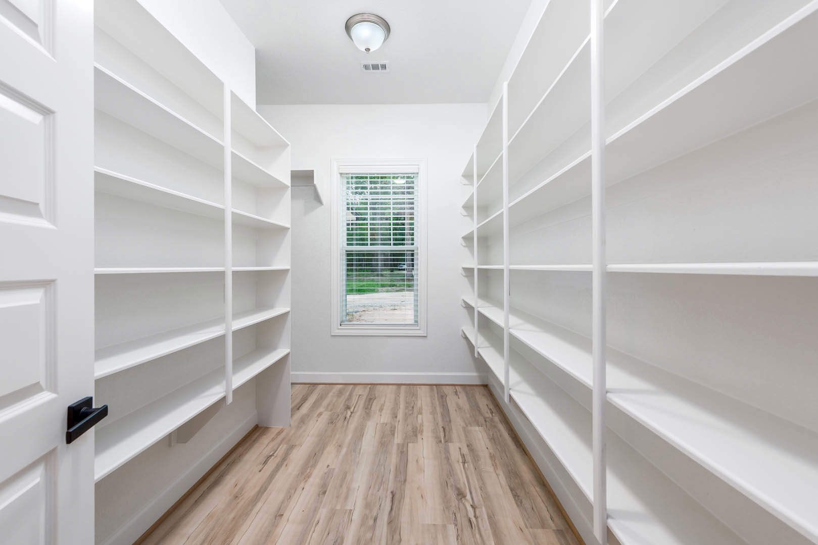 White built-in shelves along a white wall, hardwood flooring, window with blinds, ceiling light fixture, and a close-up of a brushed metal door handle.