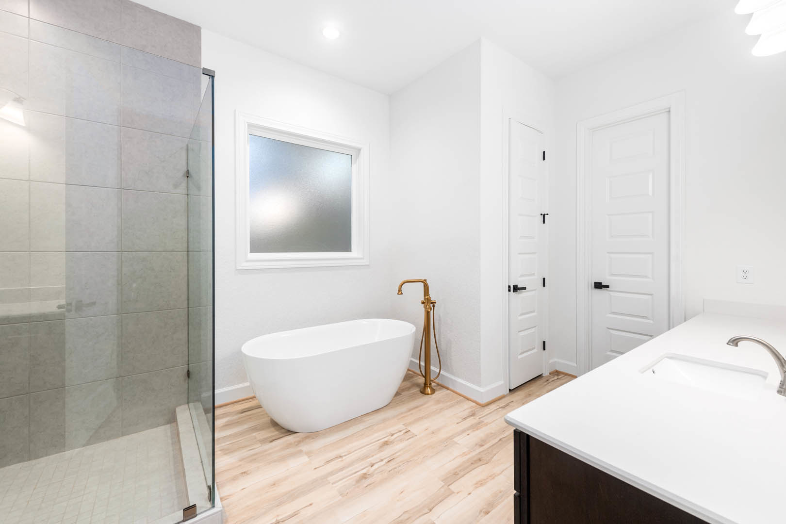 White freestanding bathtub on wood floor beside glass-enclosed shower, white countertop with chrome faucet, window with trim, and white door with black handle.