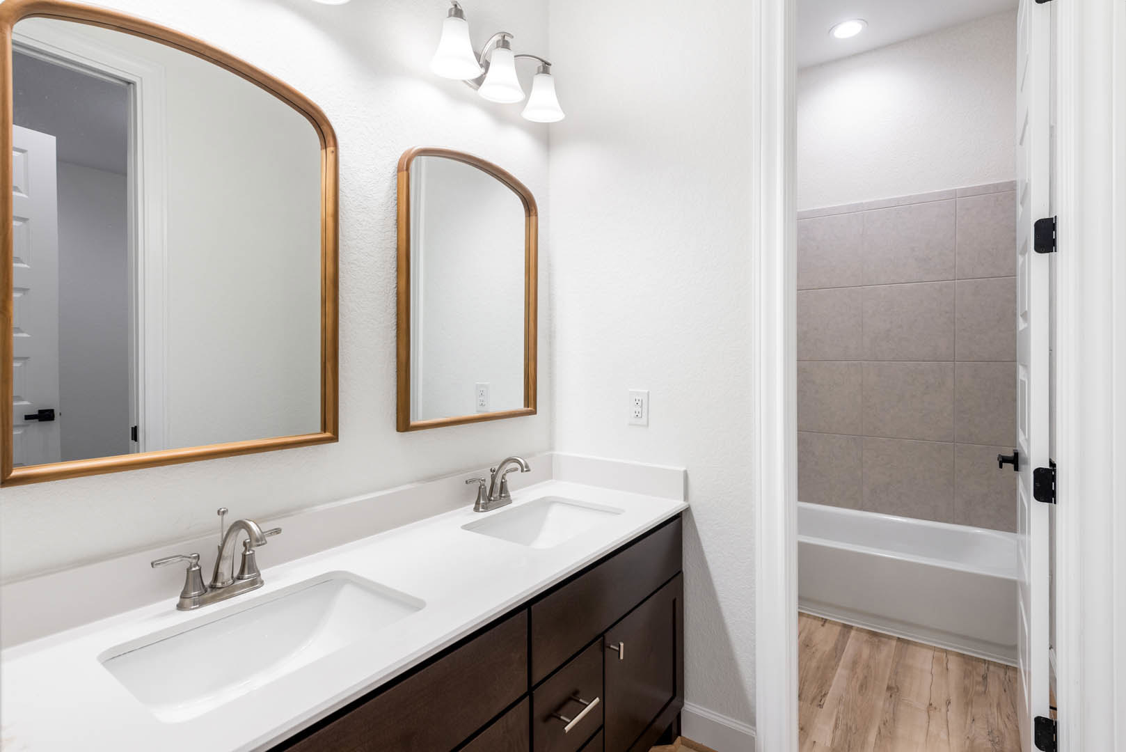 Bathroom with double sinks, freestanding bathtub, wood-framed mirror, open cabinet door, chrome faucets, tile flooring, and modern light fixtures