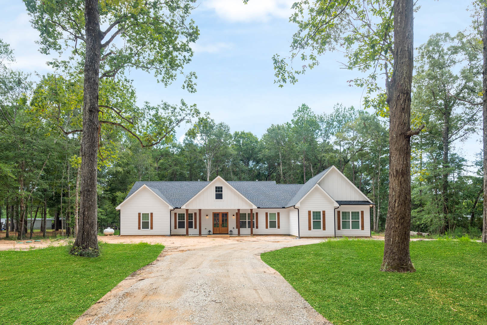 White farmhouse with double wood and glass entry doors, surrounded by mature trees, green lawn, and paved driveway under a partly cloudy sky