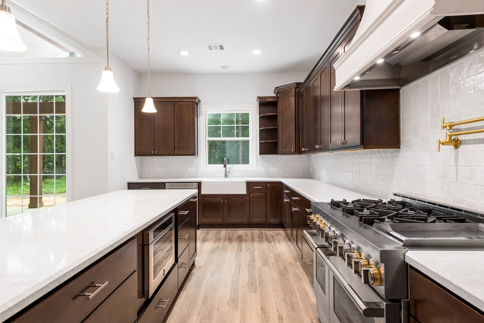 Kitchen featuring dark wood cabinetry, white countertops, stainless steel microwave, white-framed window overlooking forest, and visible plumbing pipe detail