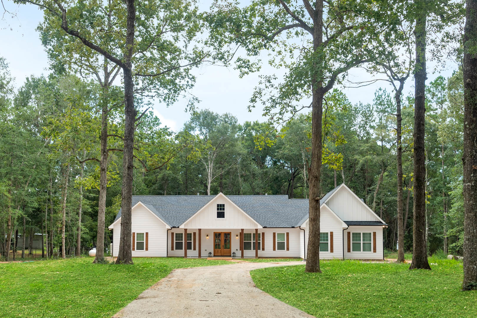 White farmhouse with black roof surrounded by mature trees, gravel path leading to front entrance, lush green lawn in foreground