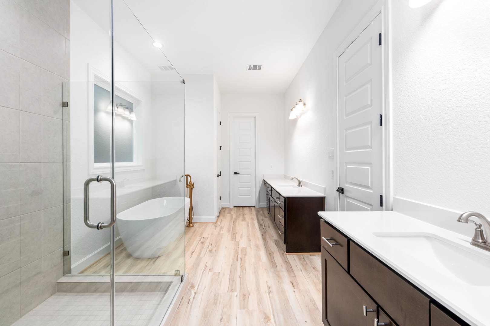 Bathroom with glass shower enclosure, freestanding white bathtub, white sink with chrome faucet, light fixture mounted on wall, white door with black handle and hinges, neutral