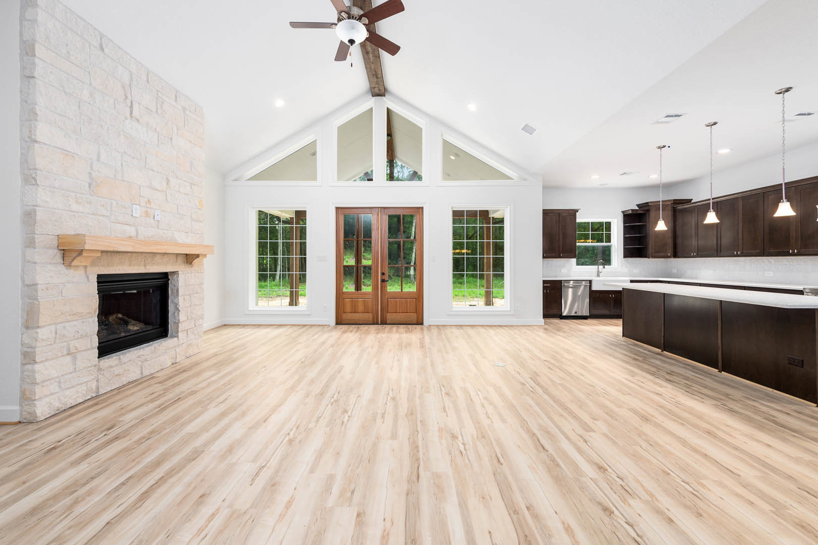 Spacious living area featuring hardwood floors, a glass-paneled double door, built-in fireplace with glass window, ceiling fan with light fixture, and partial view of kitchen