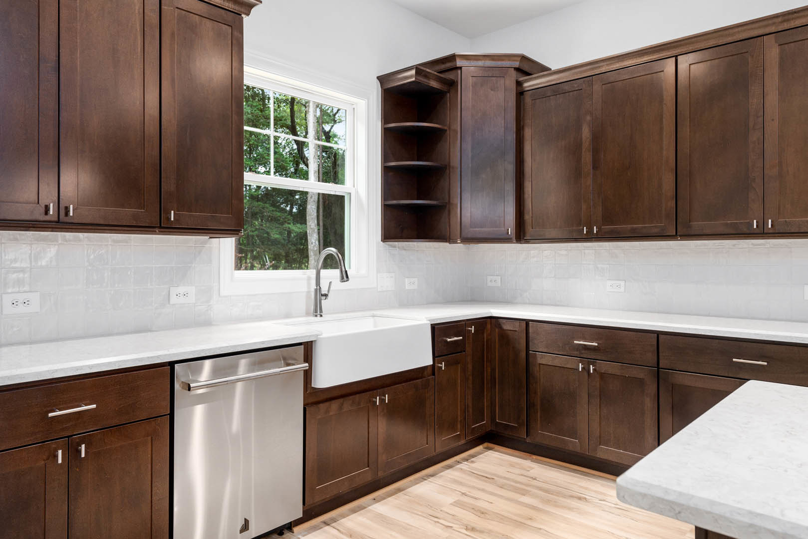 Dark wood kitchen cabinets, white granite countertop with stainless steel sink and faucet, built-in dishwasher with handle, window overlooking trees, open shelving above counter.