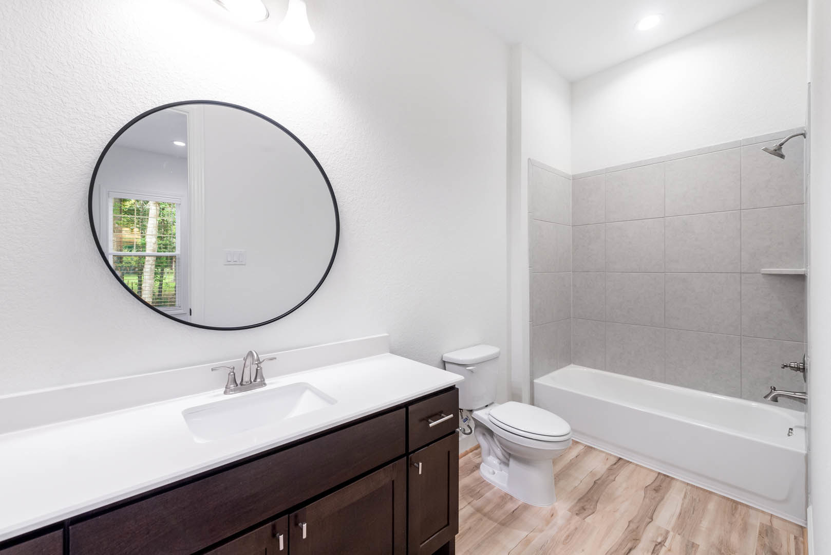 Round mirror above a white sink with chrome faucet, wood floor, white bathtub, and tiled walls in a modern bathroom.