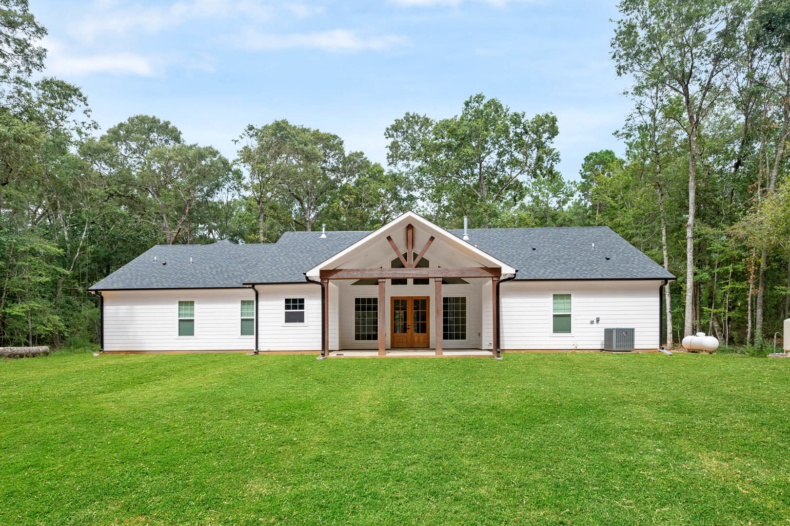 White farmhouse-style home with double glass-paneled wooden doors, grid windows, and a manicured green lawn bordered by trees under a partly cloudy sky.