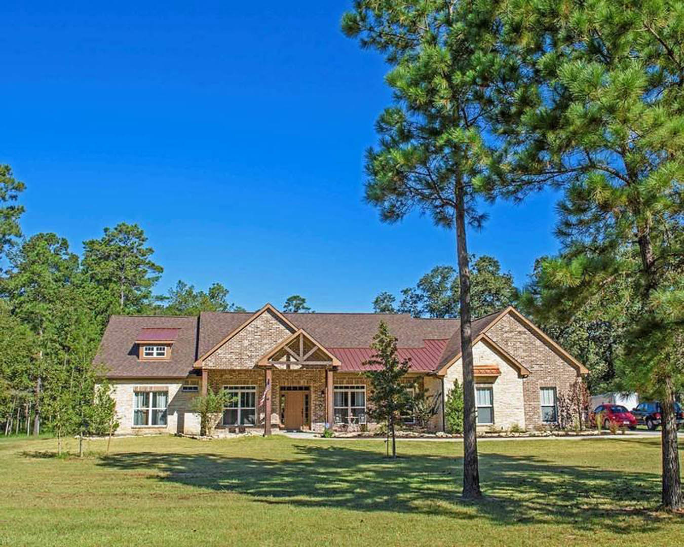 Two-story house with red roof, white siding, and large front lawn bordered by mature trees; sunlight casts tree shadows across green grass, with a red car partially visible in the
