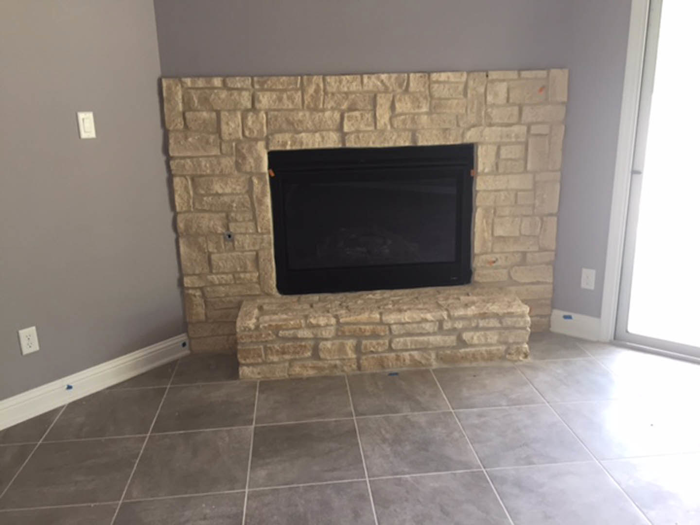 Stone fireplace with black wood-burning stove, fire screen, and hearth set in a brick wall; nearby white electrical outlet and light switch on painted wall; hardwood floor visible.