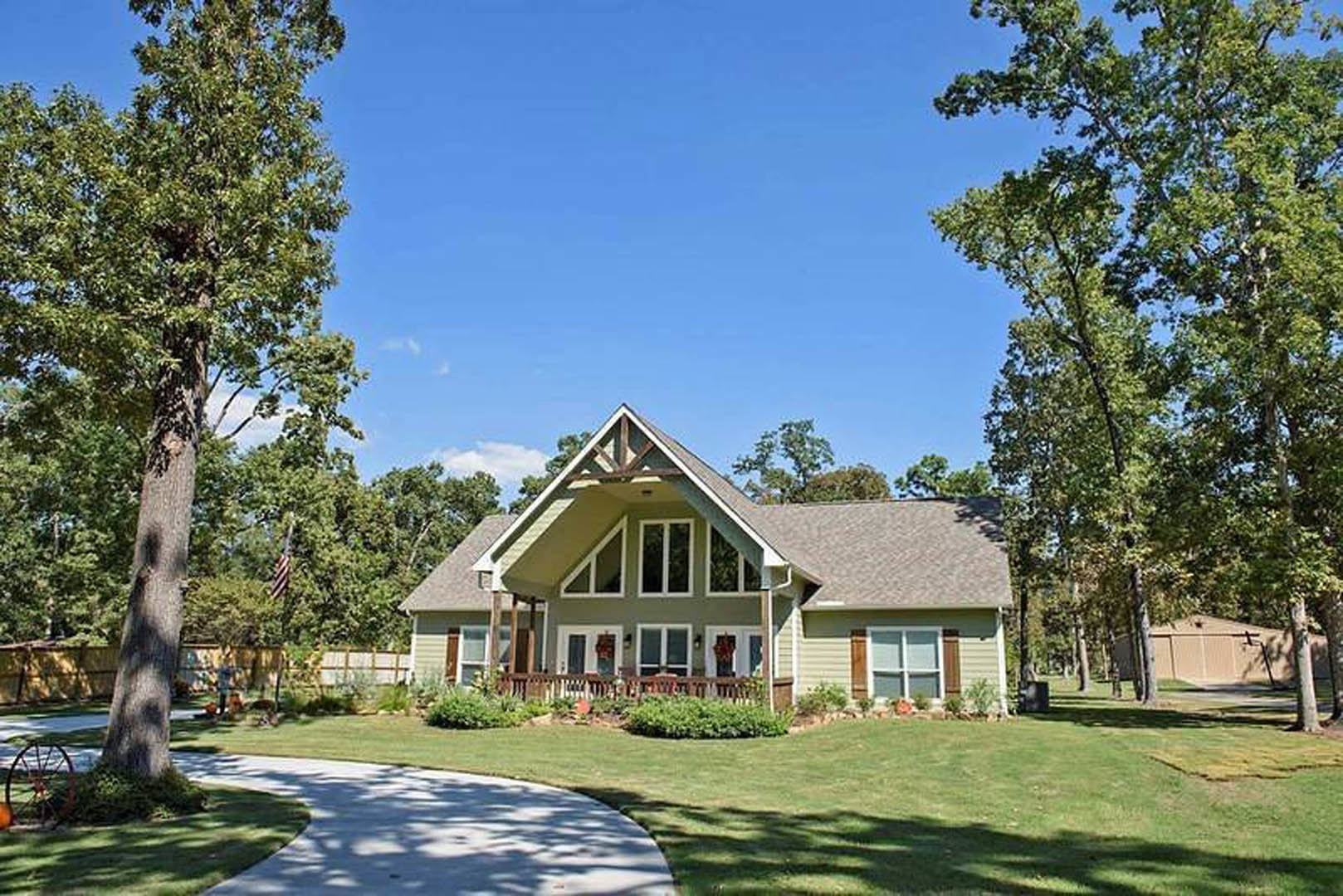 Two-story farmhouse with gabled roof, covered front porch, manicured lawn, mature trees, and American flag in the background