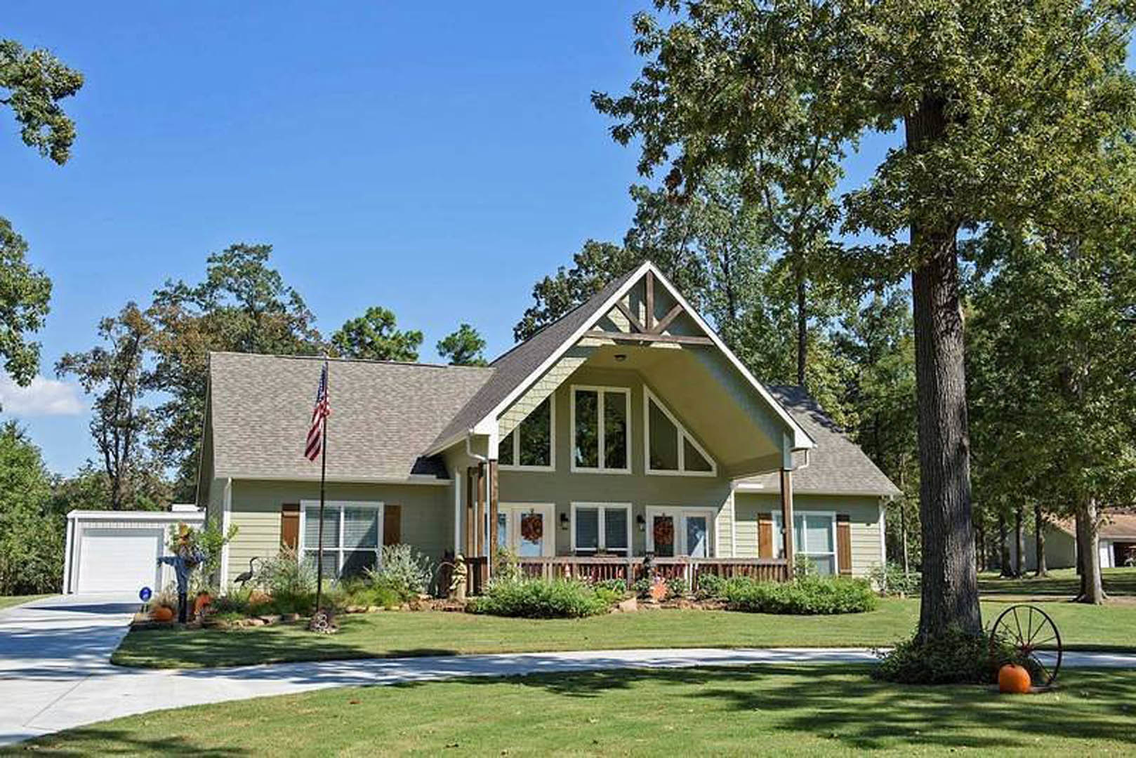 Two-story house with white siding, green front door with white trim, covered porch, American flag mounted near entrance, landscaped garden with grass, large tree in background